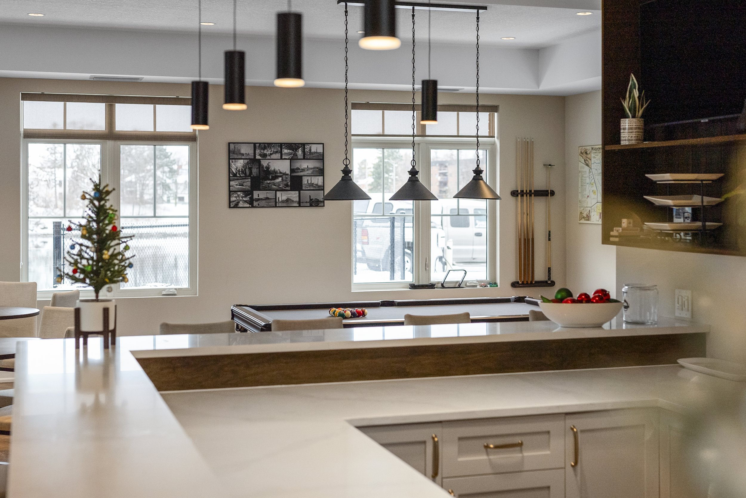 A modern kitchen and living area decorated for Christmas with a small Christmas tree on a white counter, holiday ornaments in a bowl, a pool table near the window, and framed artwork on the wall.