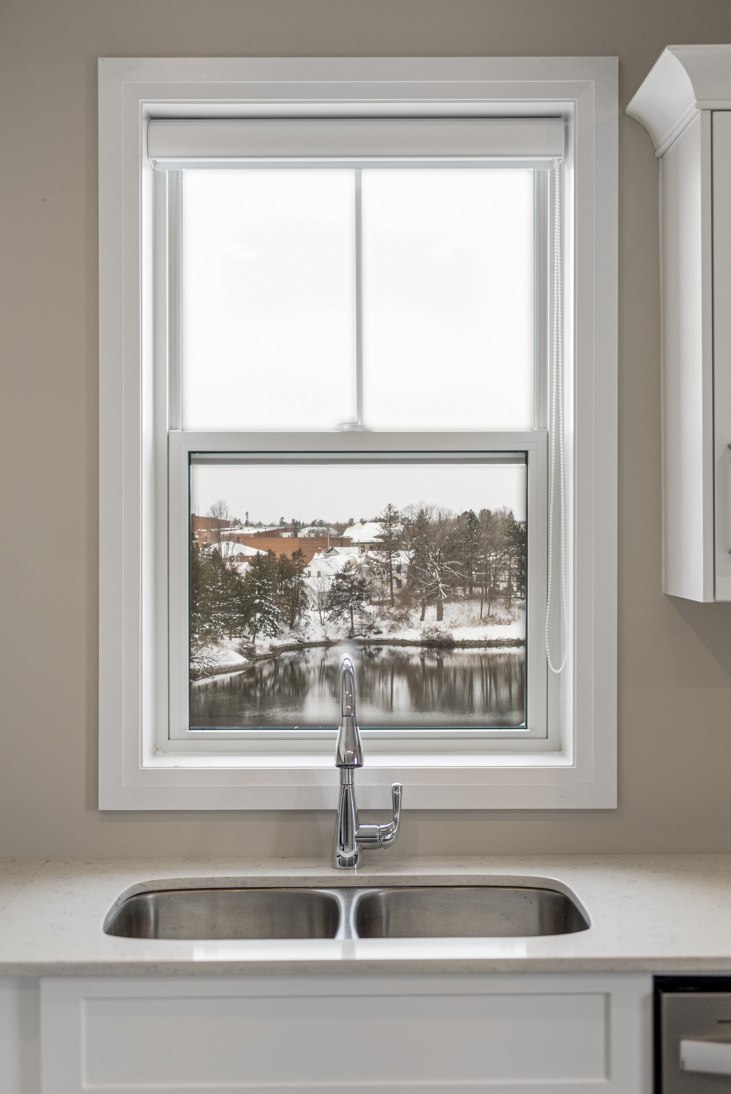 Kitchen sink with a view of a snowy riverside through a window, white window frame, and beige wall.
