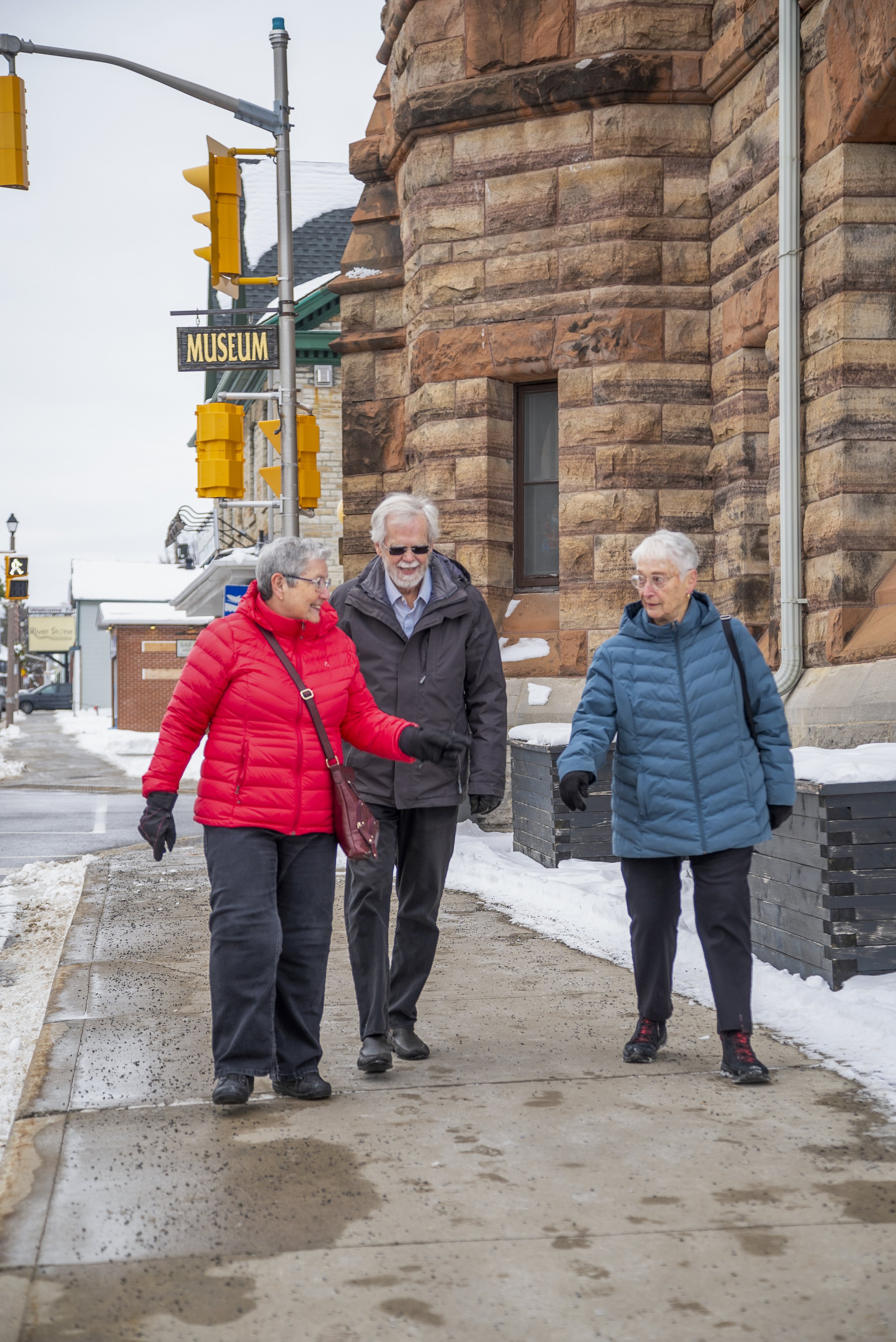 Three elderly people walking and talking on a snowy sidewalk in Arnprior, with a brown stone building and a sign reading 'Museum' in the background.