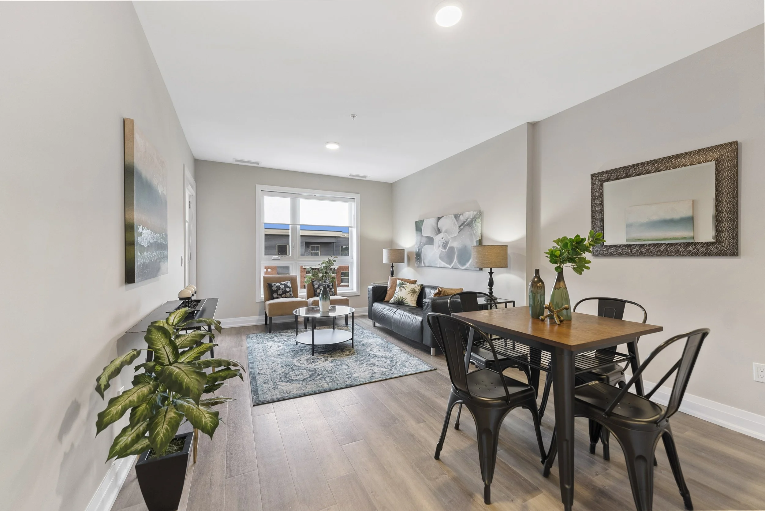 Living room with beige walls, hardwood floors, a black leather sofa with pillows, a coffee table, window with a view of neighboring buildings, a patterned rug, side tables with lamps, and a small dining table with chairs and vases.