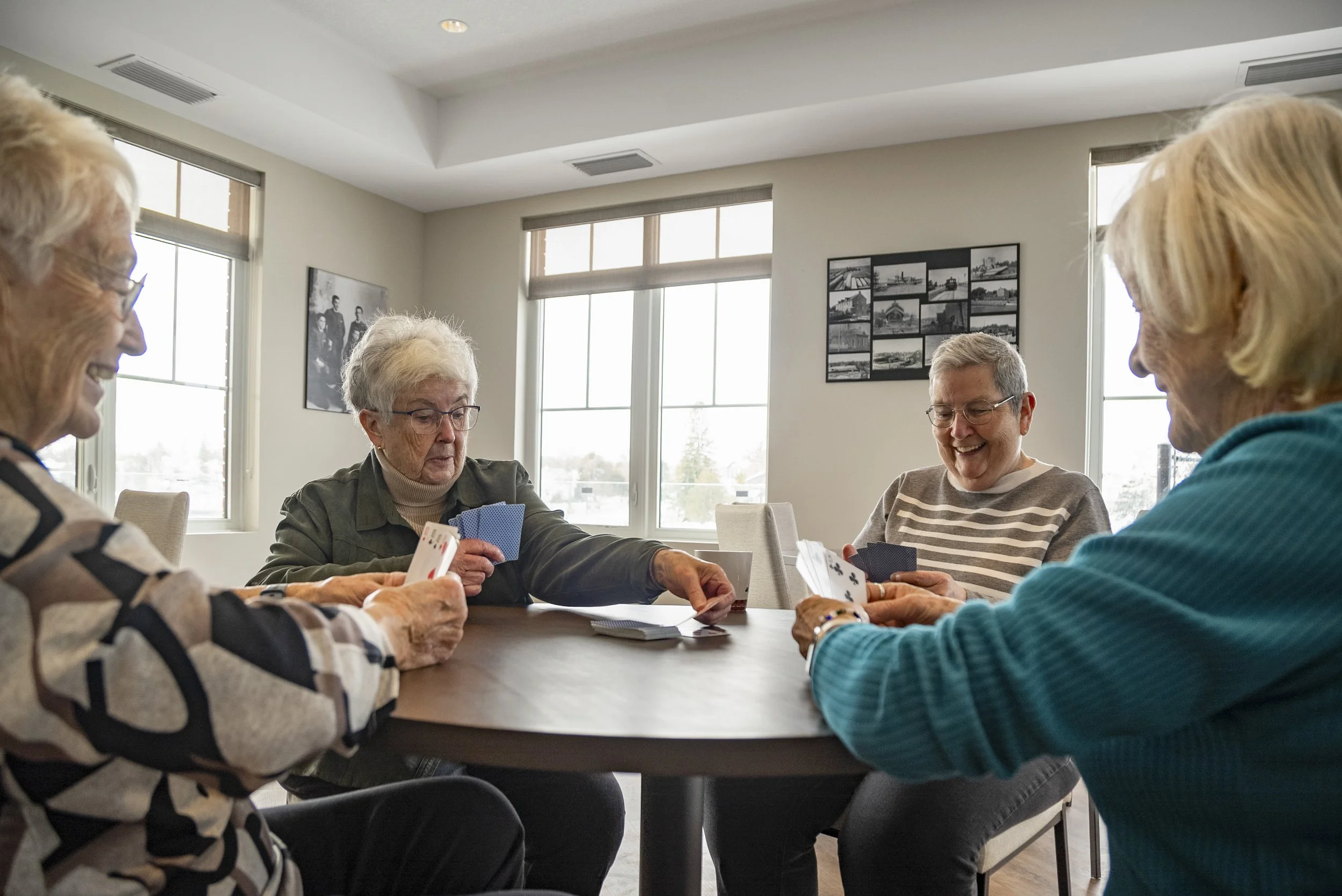 Four elderly women playing cards together at a table in a well-lit room with large windows.