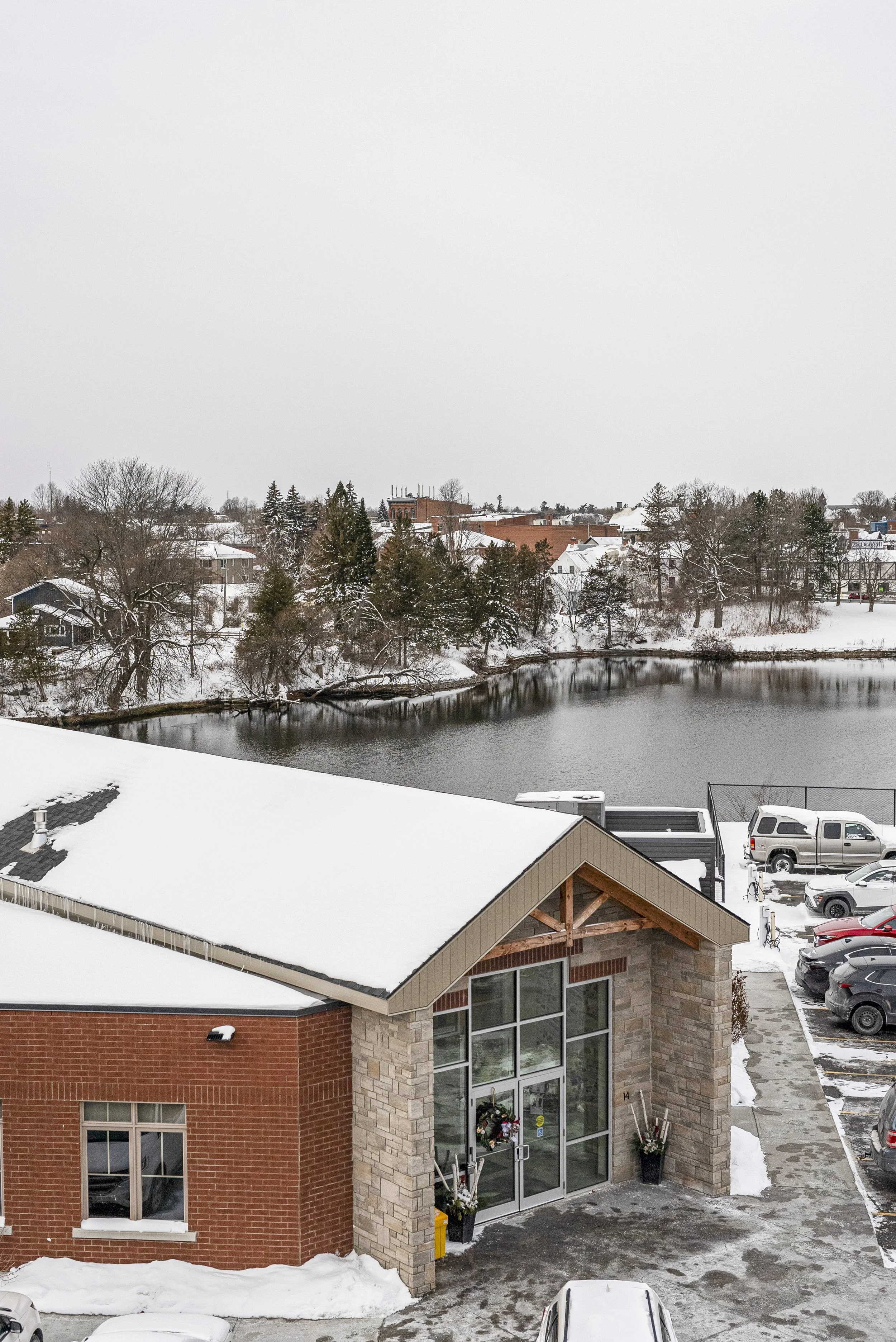 A winter scene with snow-covered rooftops and parking lot in front of a brick building with large glass doors, overlooking a partially frozen pond with snow-covered trees and houses in the background.