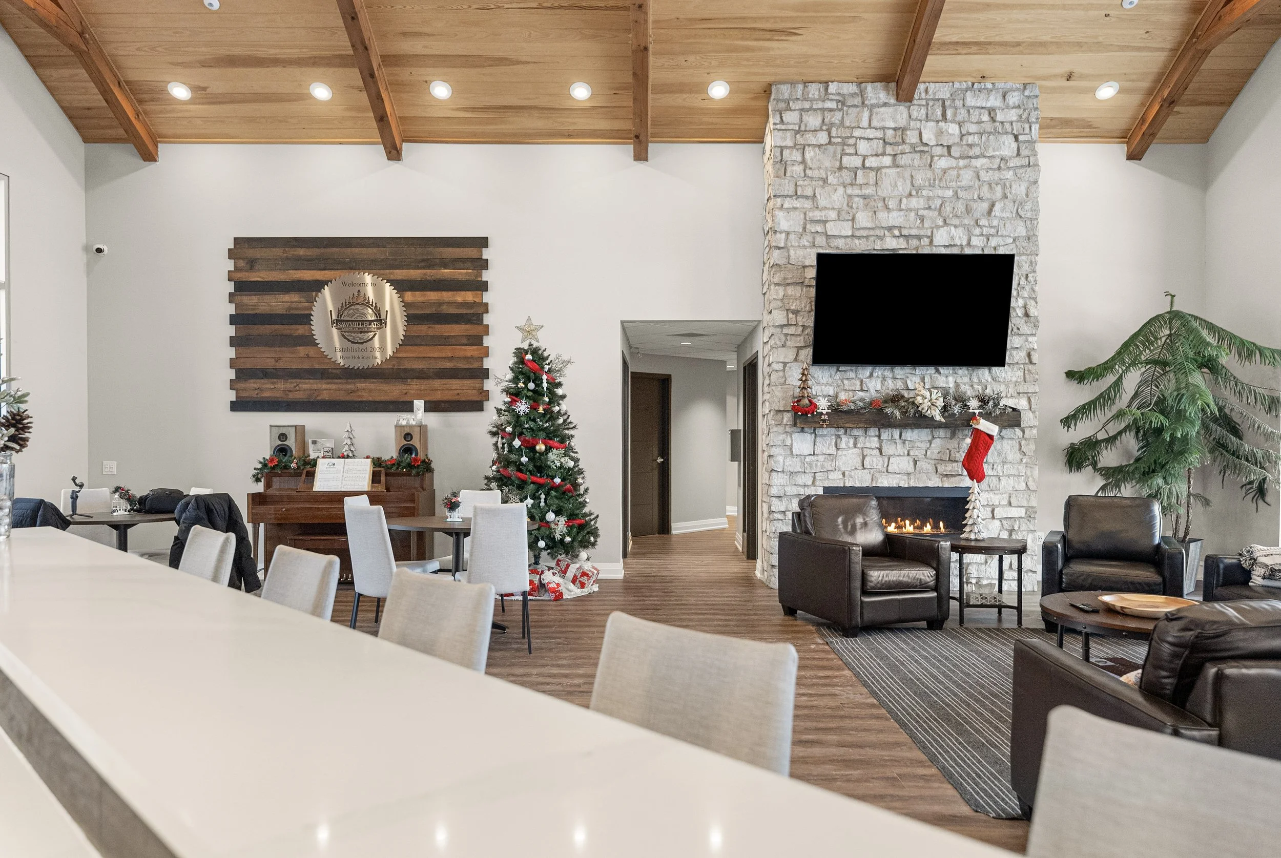 A cozy, modern living room decorated for Christmas, featuring a Christmas tree with ornaments and red ribbon, a white stone fireplace with a mantel decorated with holiday garlands, a large flat-screen TV mounted above, a black leather armchair, a lar
