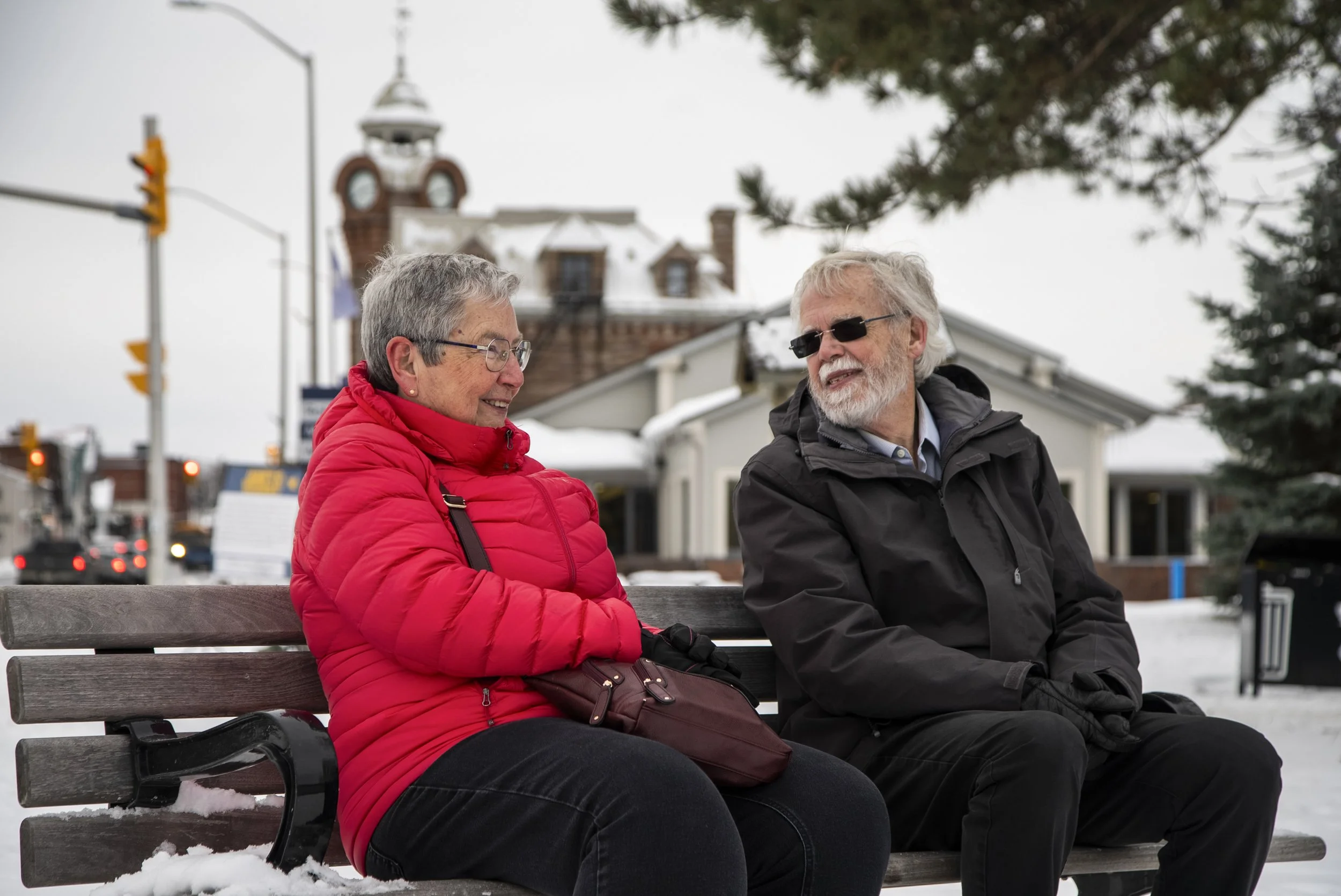 An elderly couple sitting on a park bench outdoors in winter, smiling and talking with each other, with snow on the ground and buildings in the background.