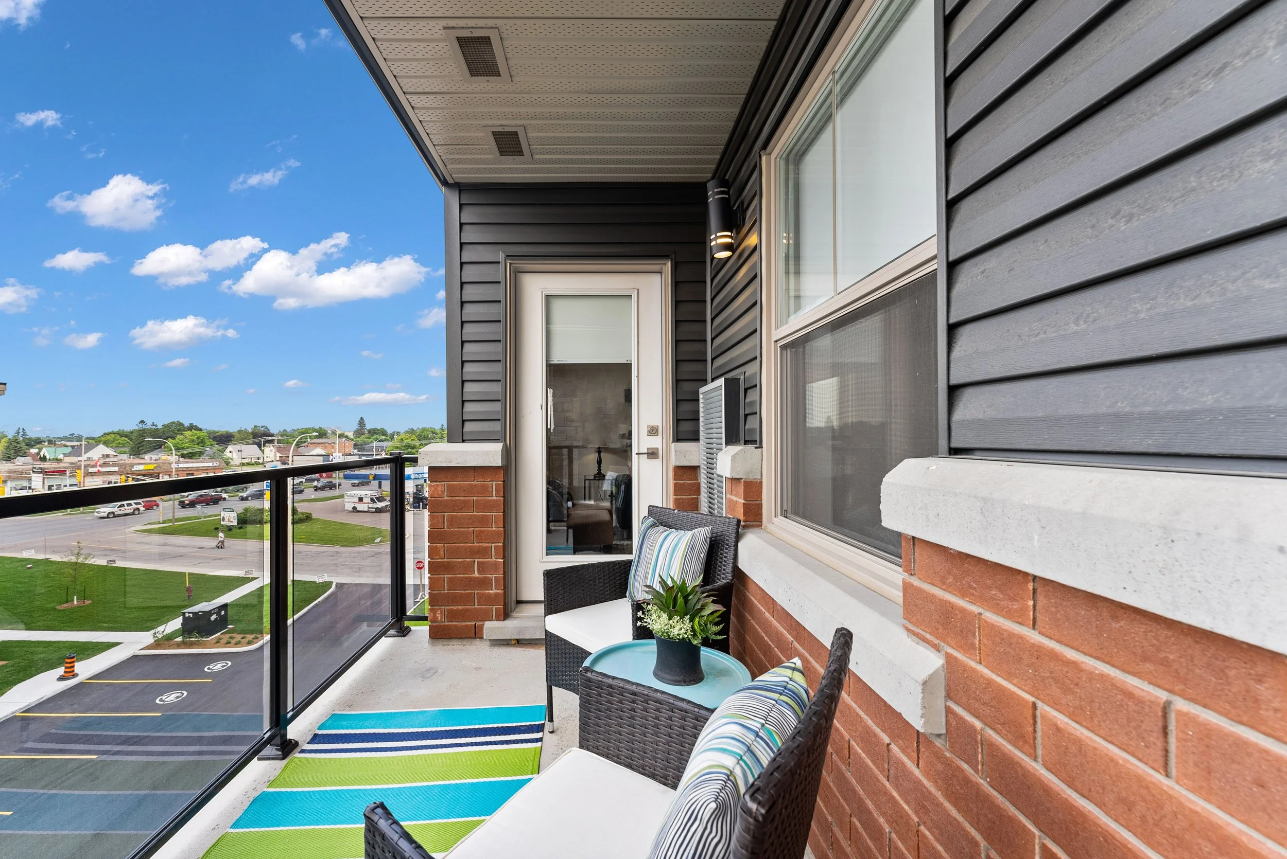 Balcony with patio furniture, small round table, chairs, potted plant, colorful striped rugs, view of parking lot and cityscape in the background.