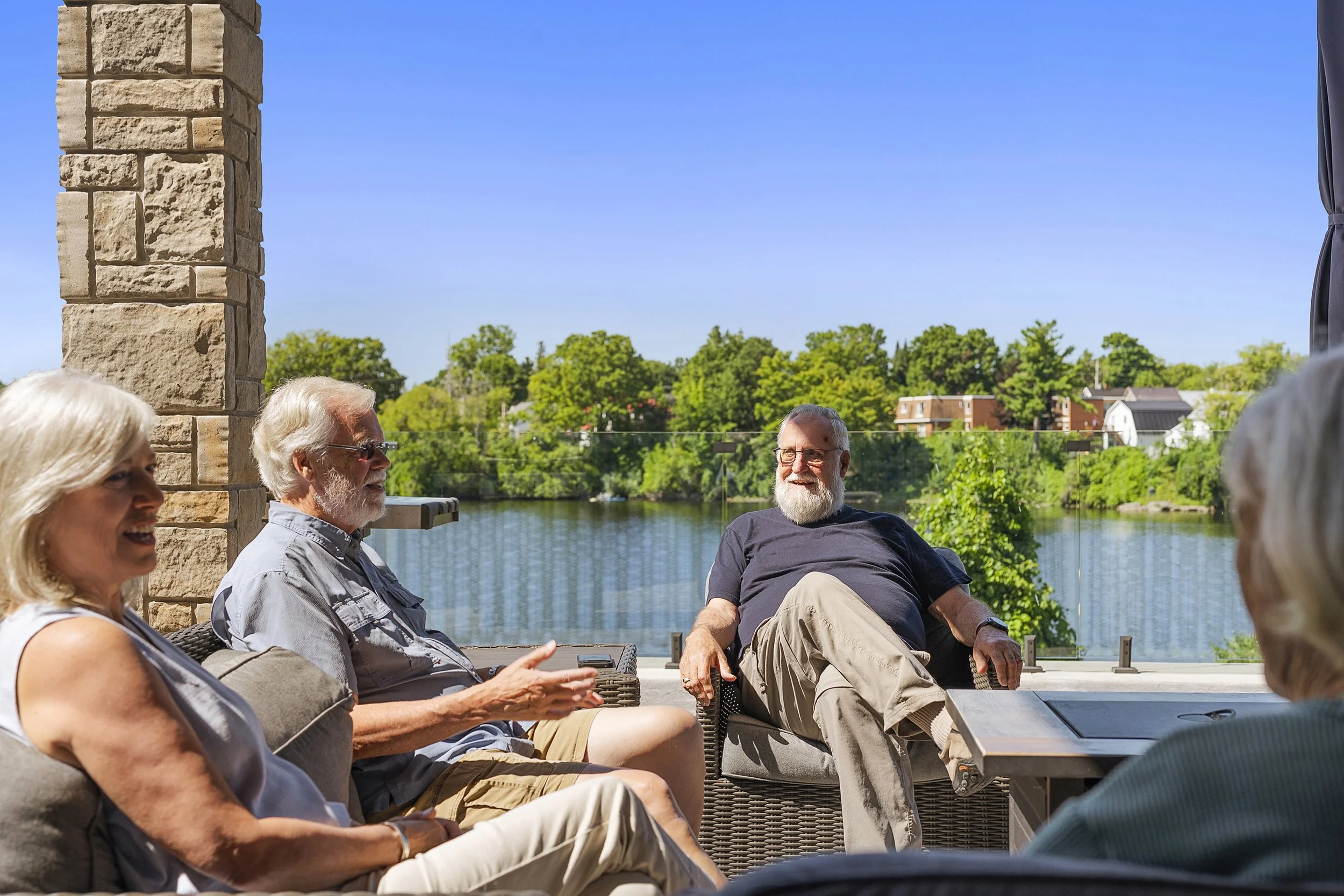 A group of older adults relaxing and chatting on outdoor patio furniture with a river and trees in the background
