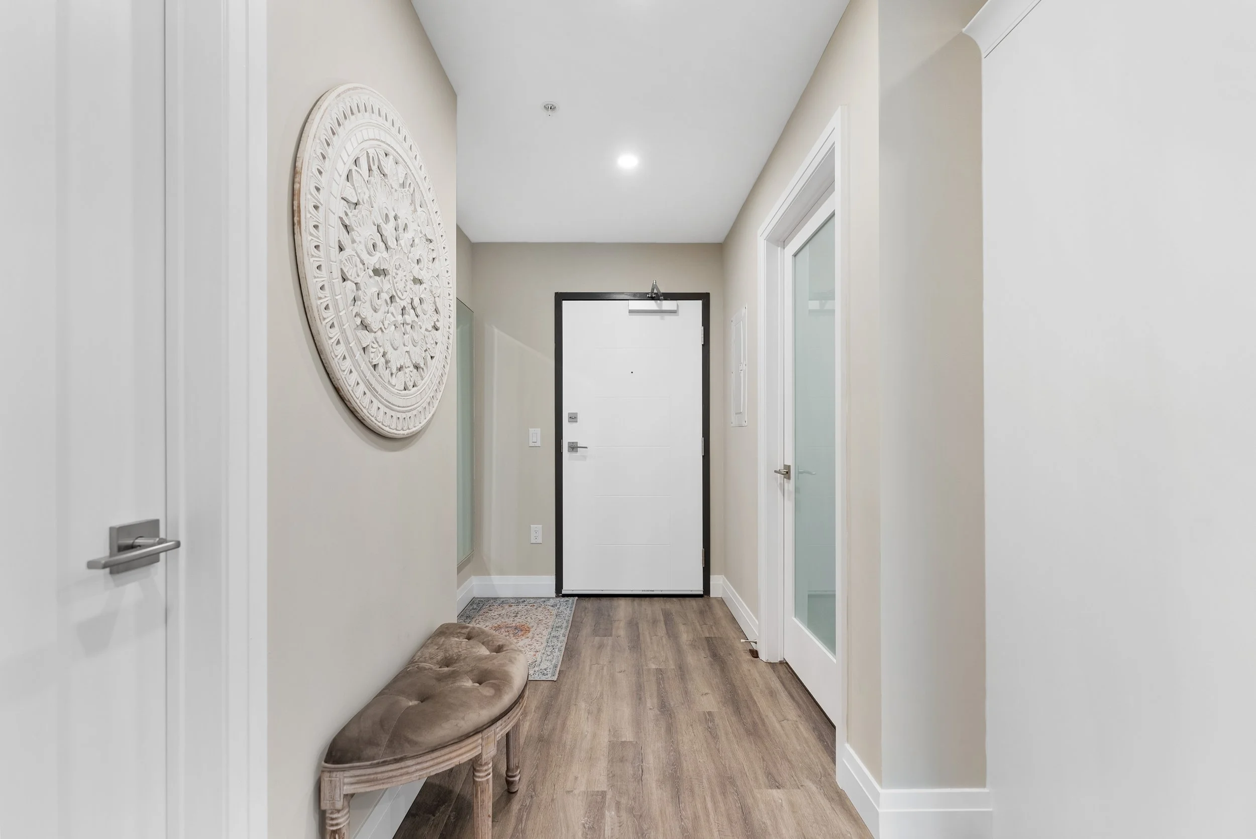 A modern hallway with beige walls, a white door at the end, and wooden flooring. There is a beige tufted bench and decorative wall art.