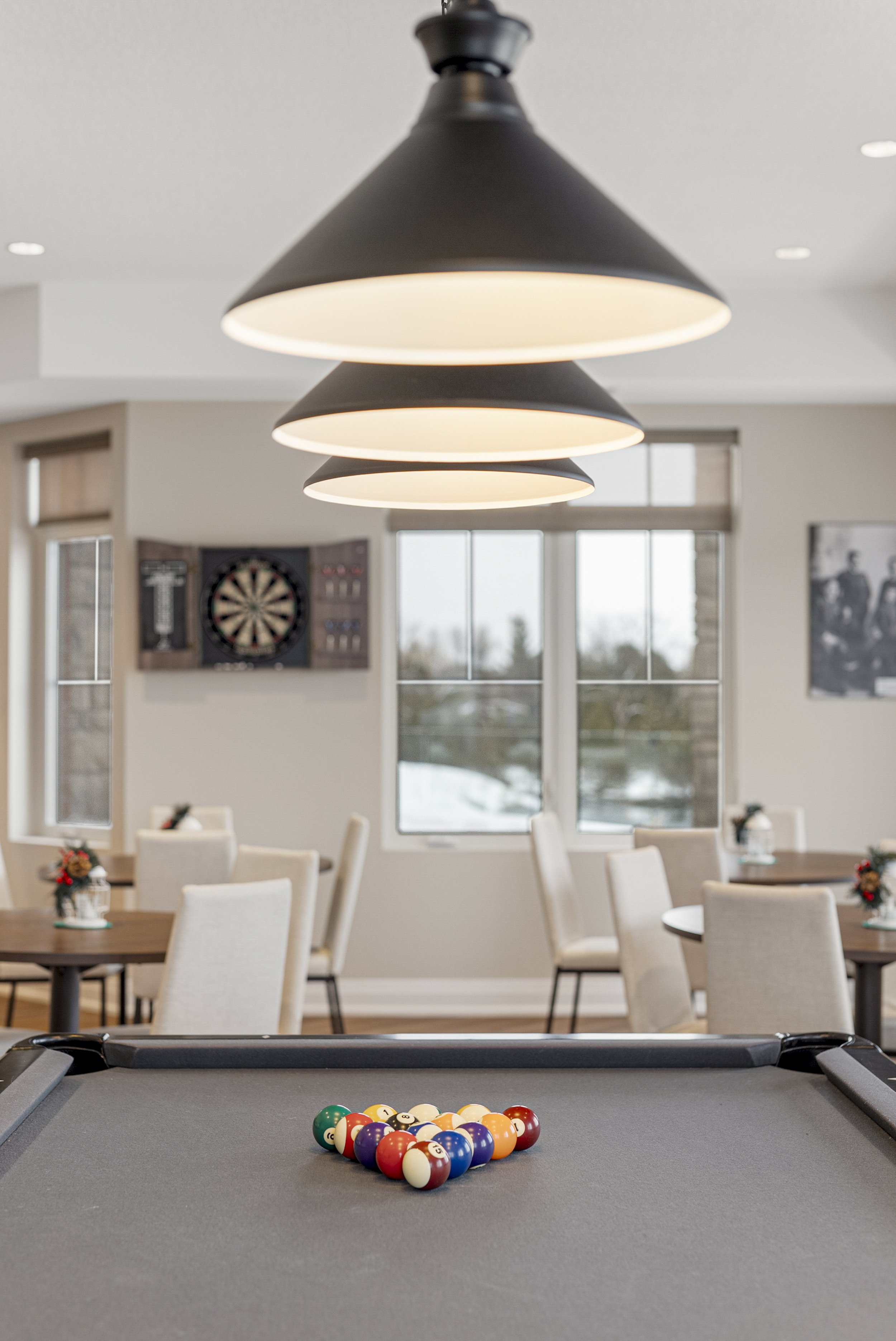 Pool table with cue balls set up in a game, in a room with hanging lights, chairs, tables, a dartboard, and windows.