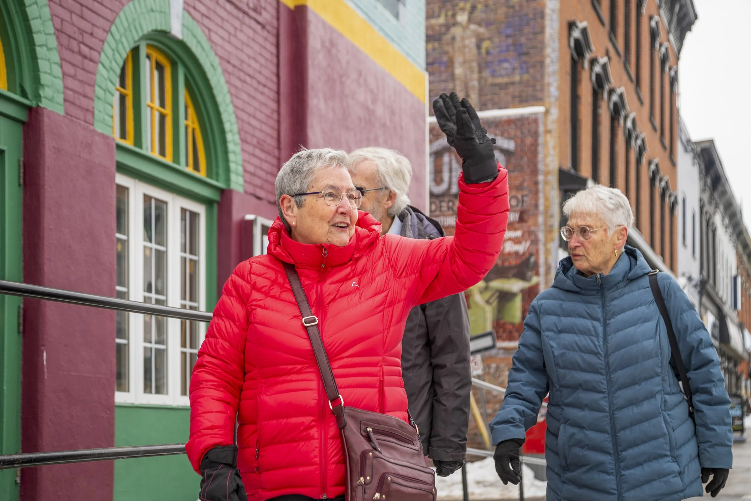 Three elderly women walking on a colorful city street, with two of them wearing winter jackets and one waving.