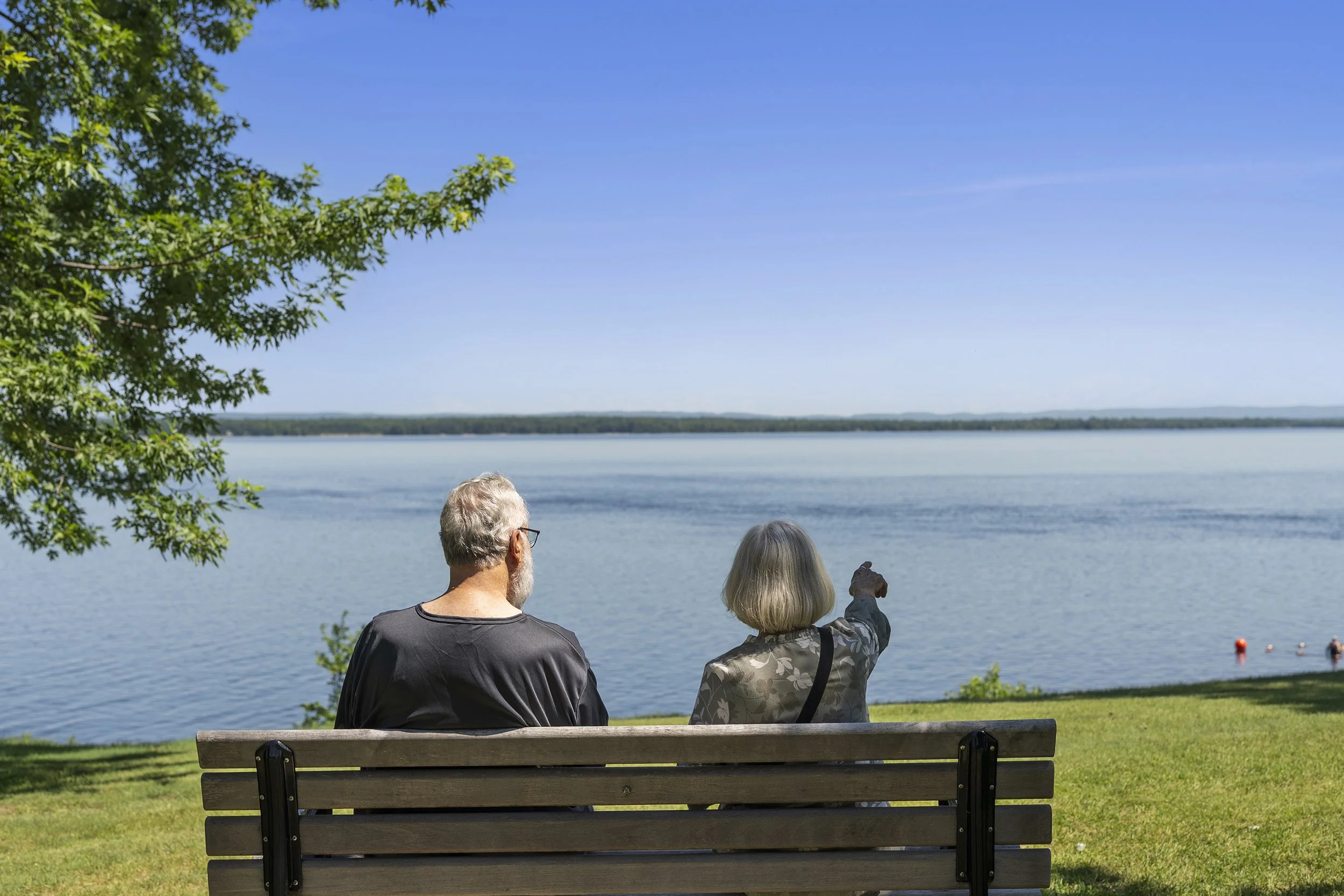 A man and woman sitting on a wooden park bench, viewed from behind, as they look out over a wide, calm body of water with one pointing across the horizon