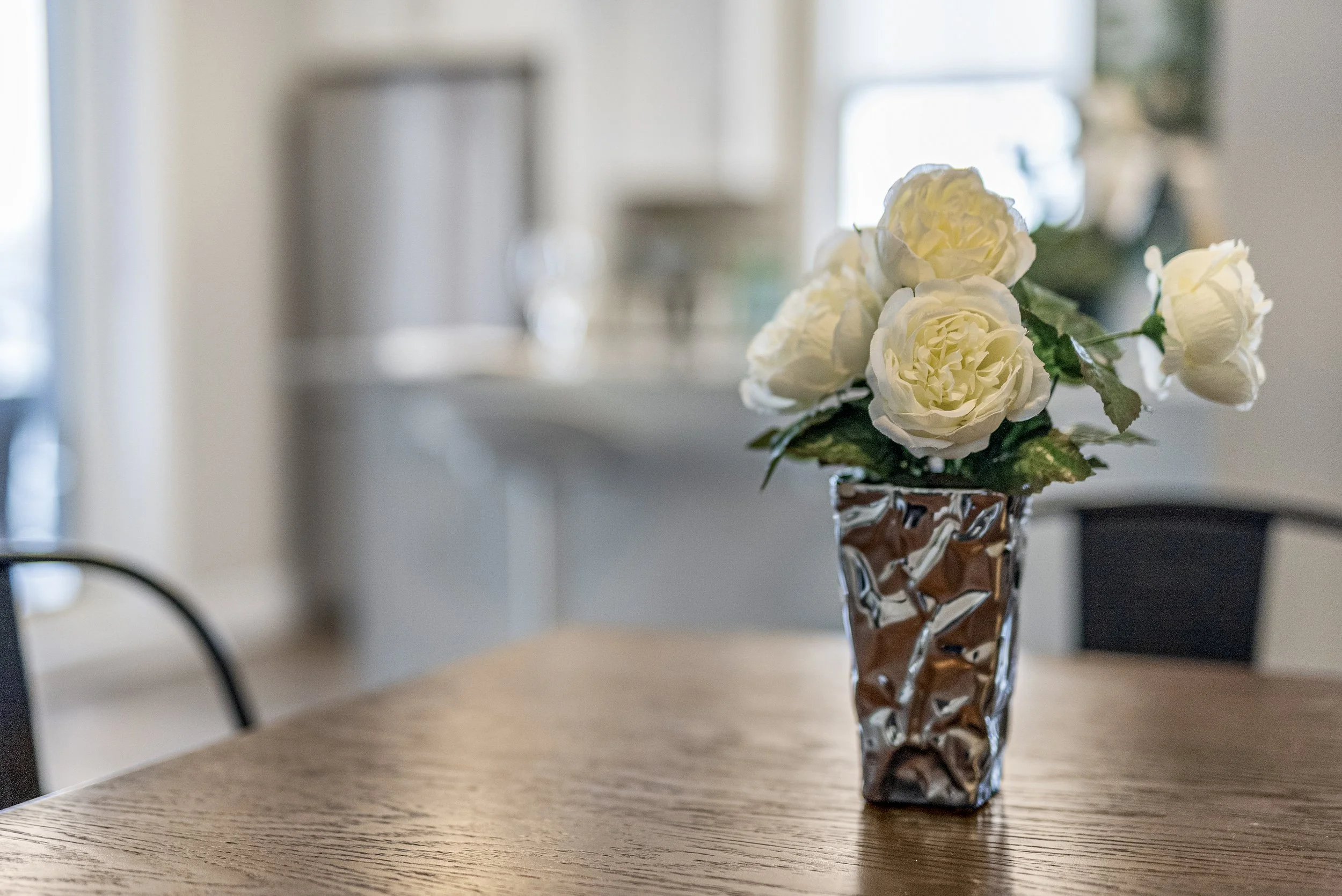 A bouquet of white flowers in a crumpled metallic silver vase on a wooden table.
