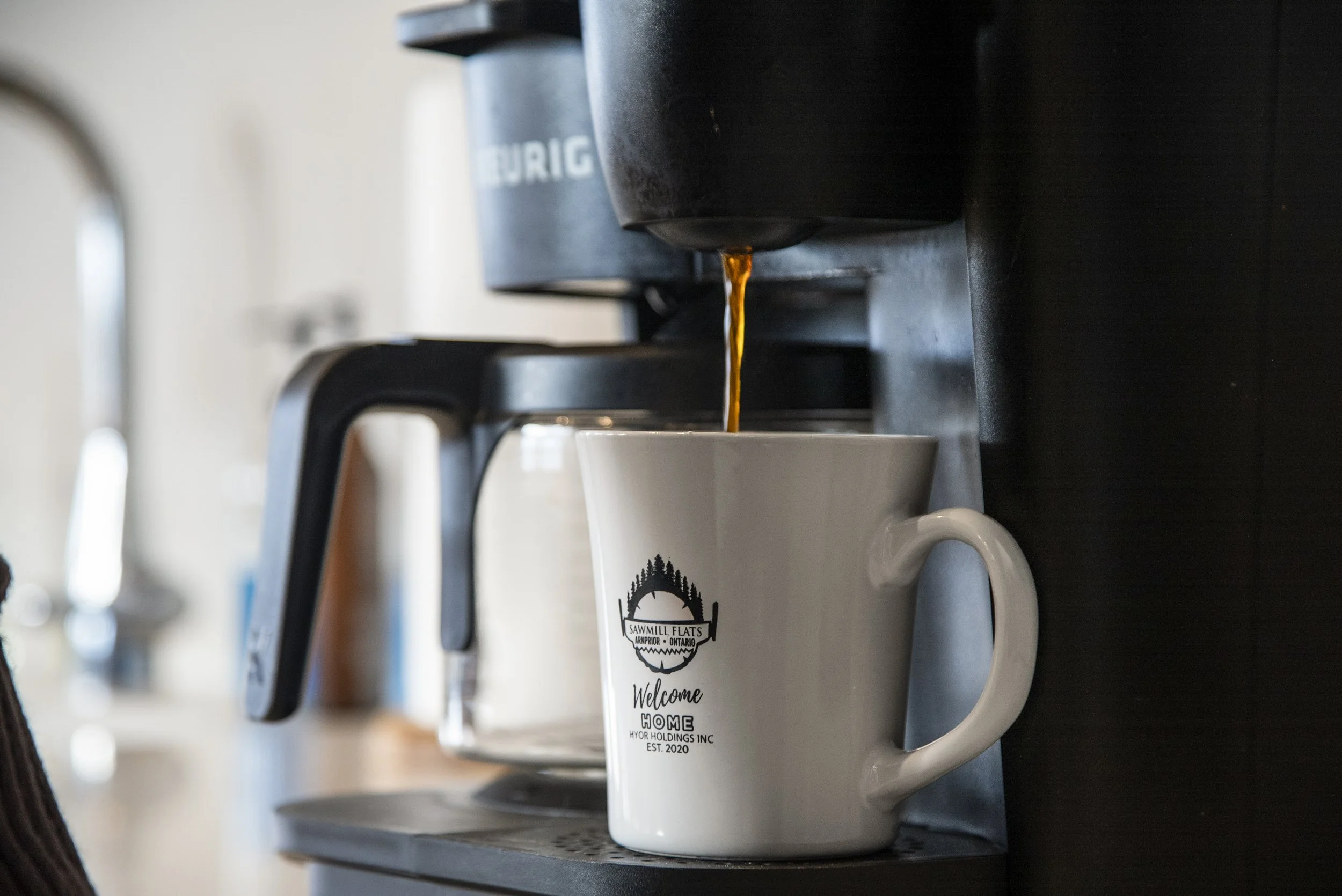 A coffee machine pouring coffee into a white mug with a logo and text that reads "Sawmill Flats, Welcome Home, Hyor Holdings Inc, Est. 2020".