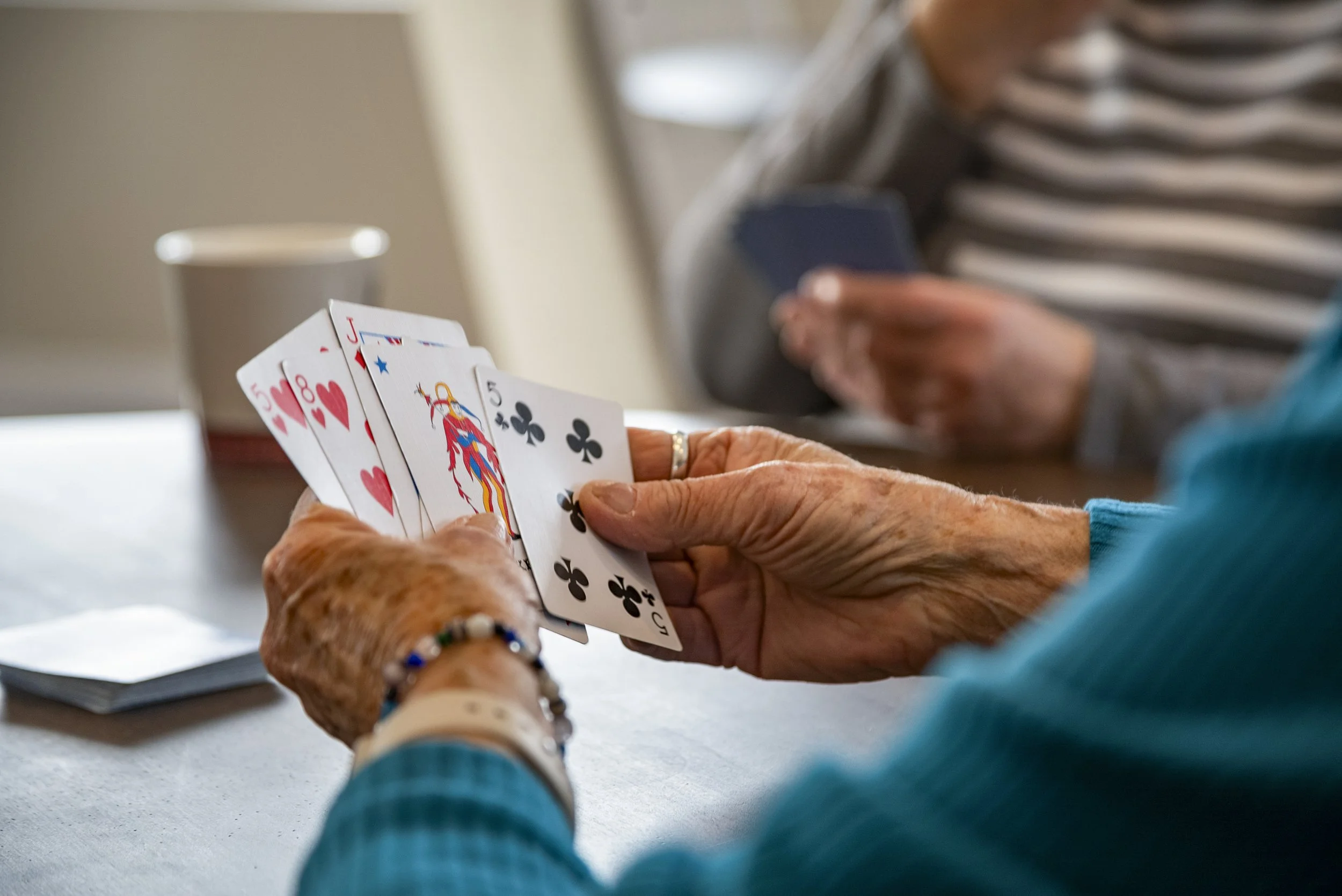 Close-up of elderly person holding a hand of playing cards, with another person in the background looking at their phone at a table.