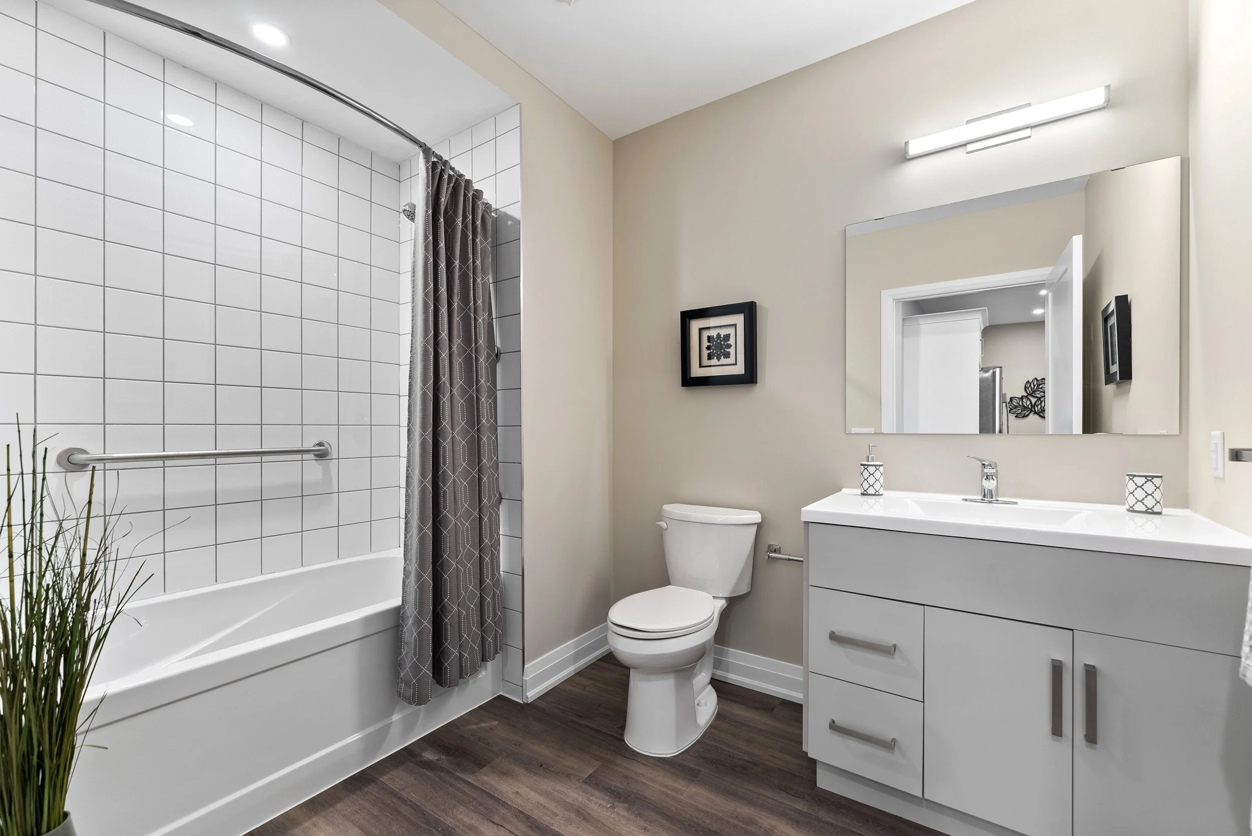 Modern bathroom with white bathtub, gray shower curtain, white toilet, white vanity with sink, large wall mirror, and wood-look flooring.