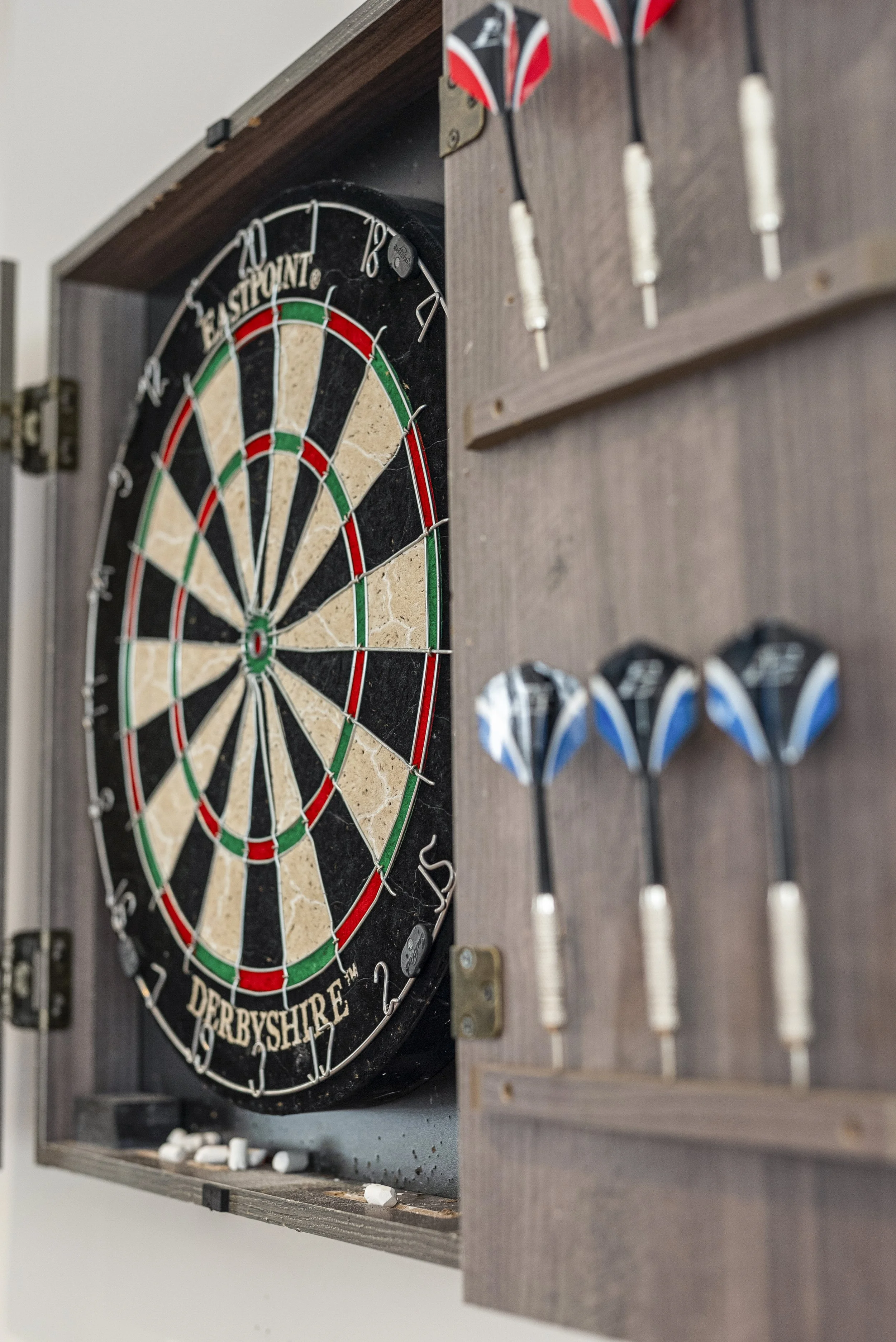 A dartboard inside a wooden cabinet with darts hanging on the side. The dartboard has black, white, red, and green sections.