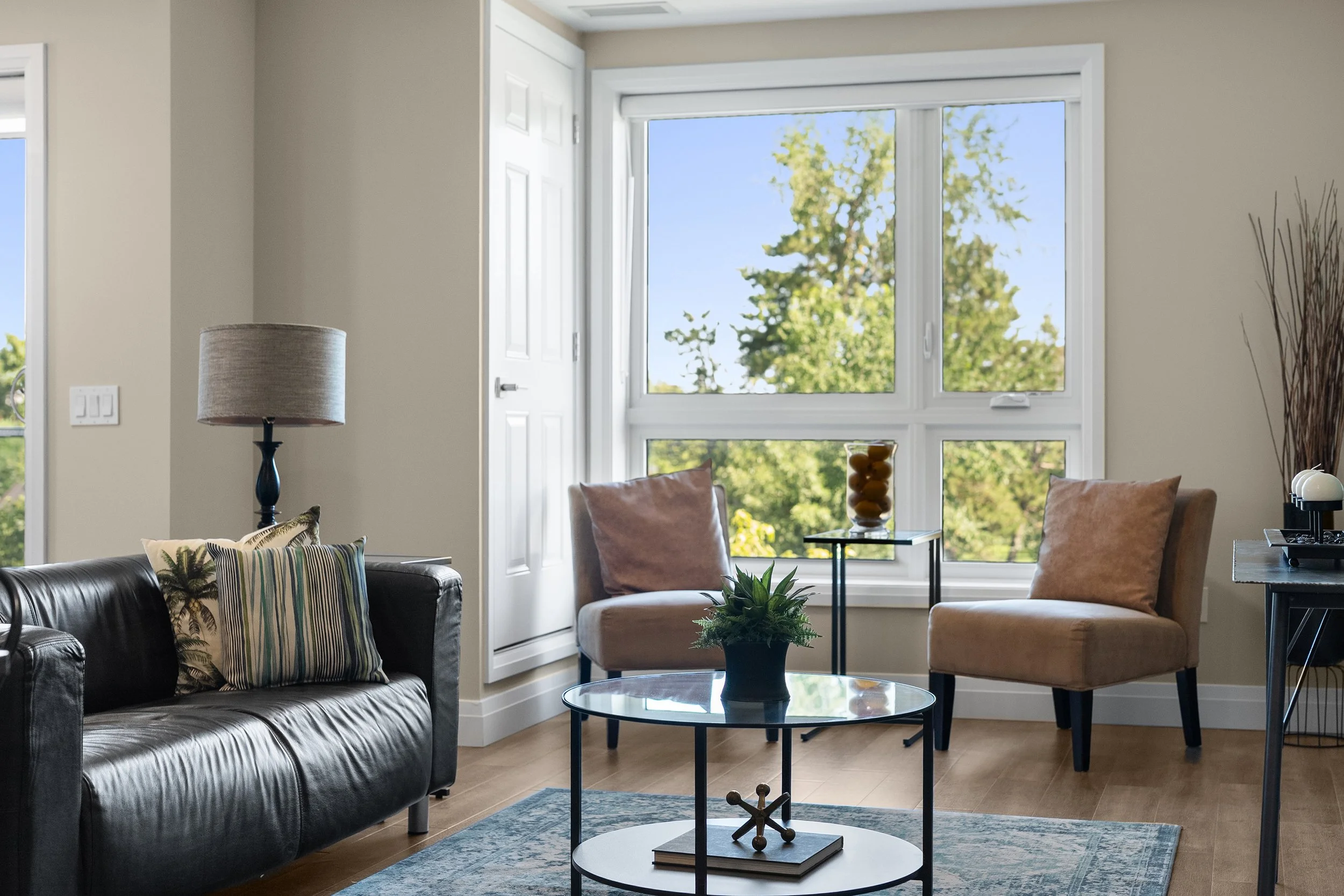 A bright, modern living room set up with a dark leather sofa, a round glass coffee table, and cozy armchair seating by a large window overlooking greenery