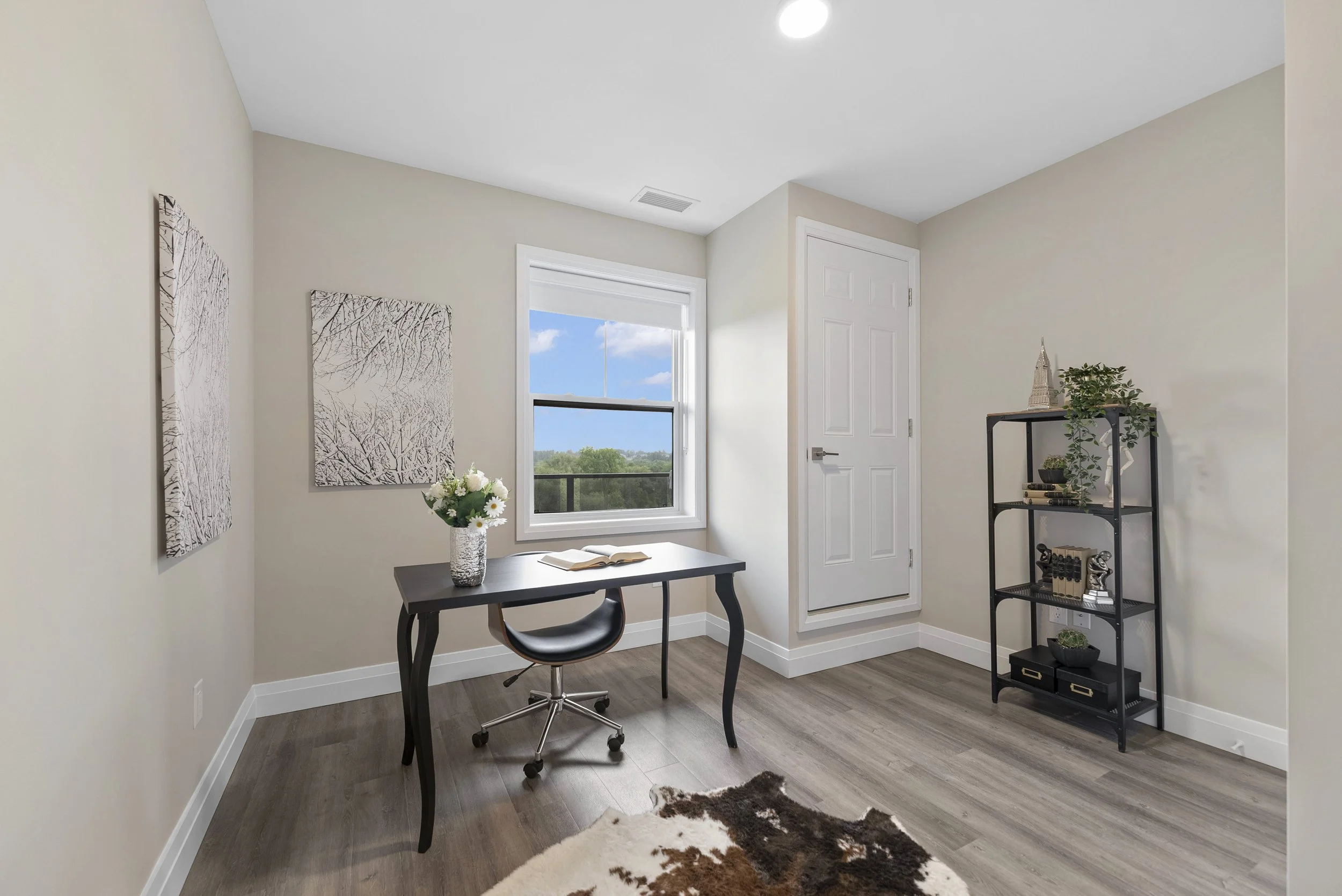 Bright home office with a black desk, gray chair, window with a view of trees, beige walls, wooden flooring, and a black metal bookshelf with decor.