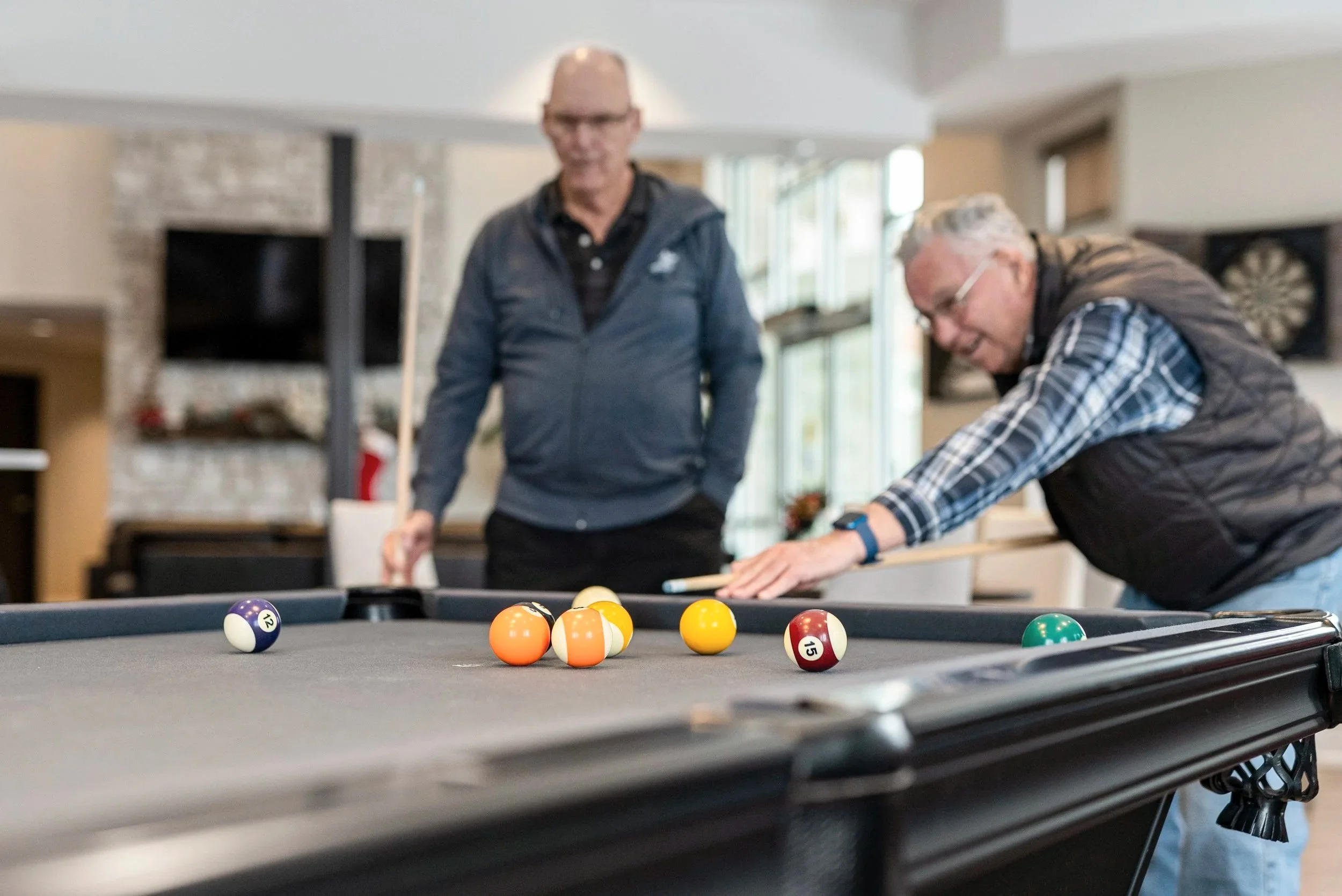 Two older men playing pool indoors, with one lining up a shot while the other observes.