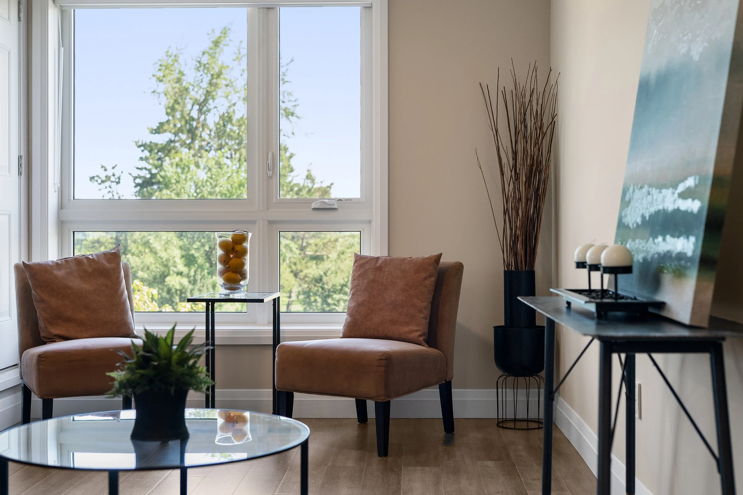 Two tan armchairs, side tables, and decorative decor in a brightly lit room with a large window looking out at trees.