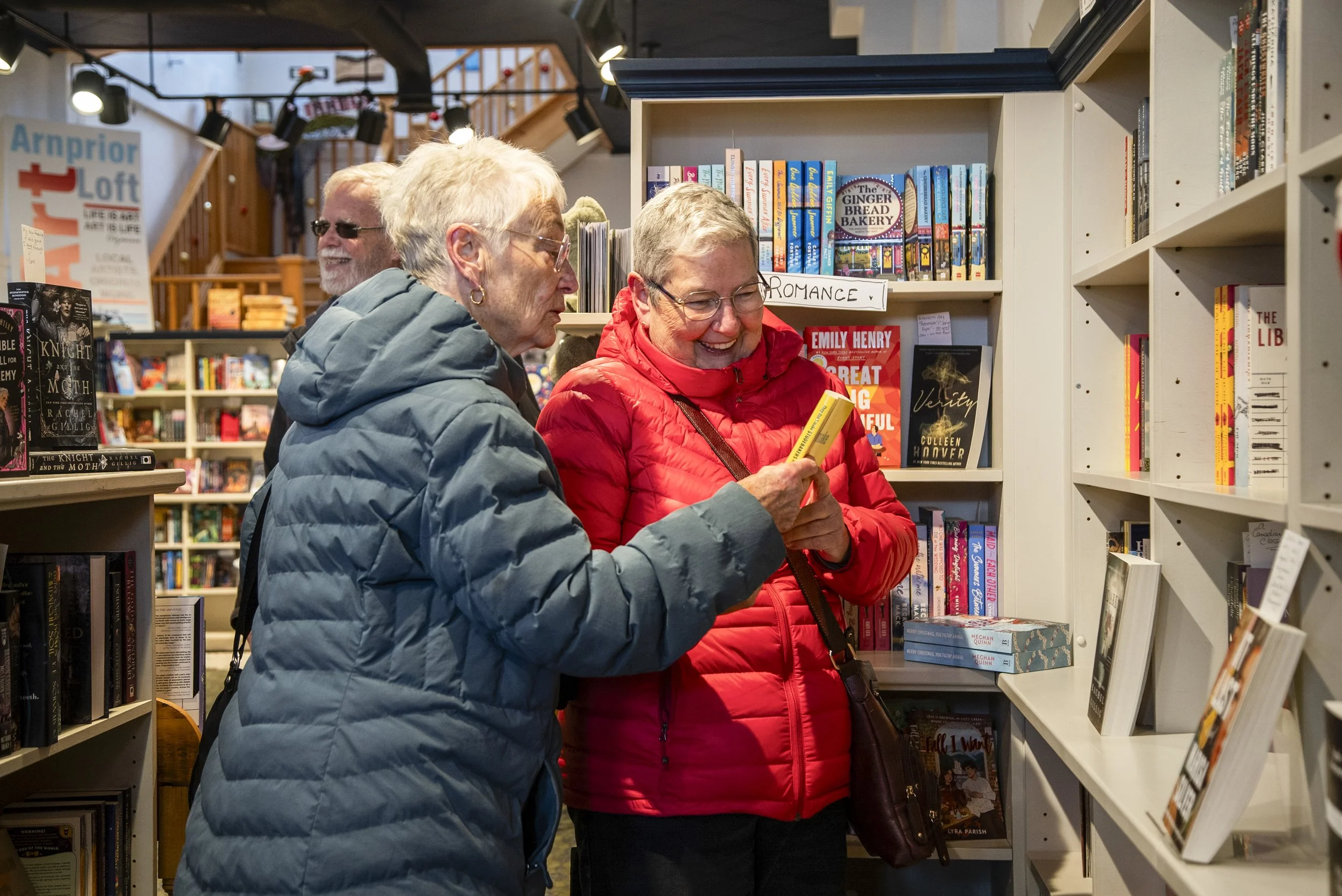 Three elderly women shopping for books at a bookstore, one wearing a light blue jacket, another in a red jacket, and the third in a black jacket, engaged in conversation and browsing shelves filled with books.