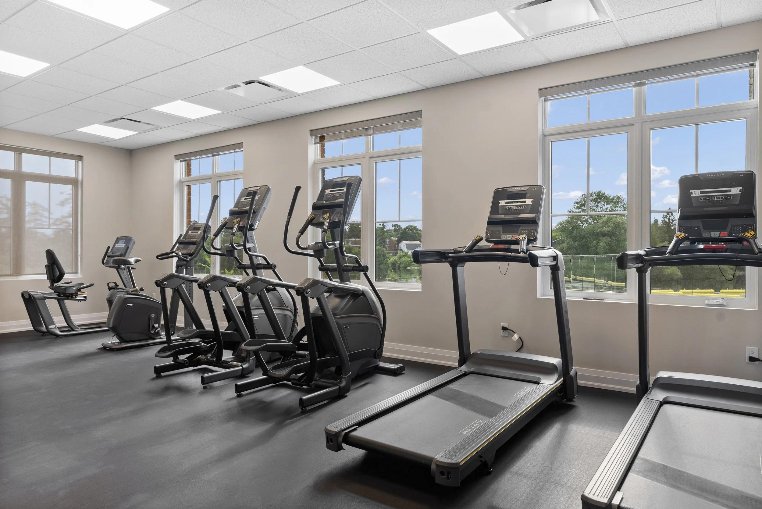 Gym with cardio equipment including treadmills, elliptical machines, and stationary bike, near large windows with a view of trees and houses outside.