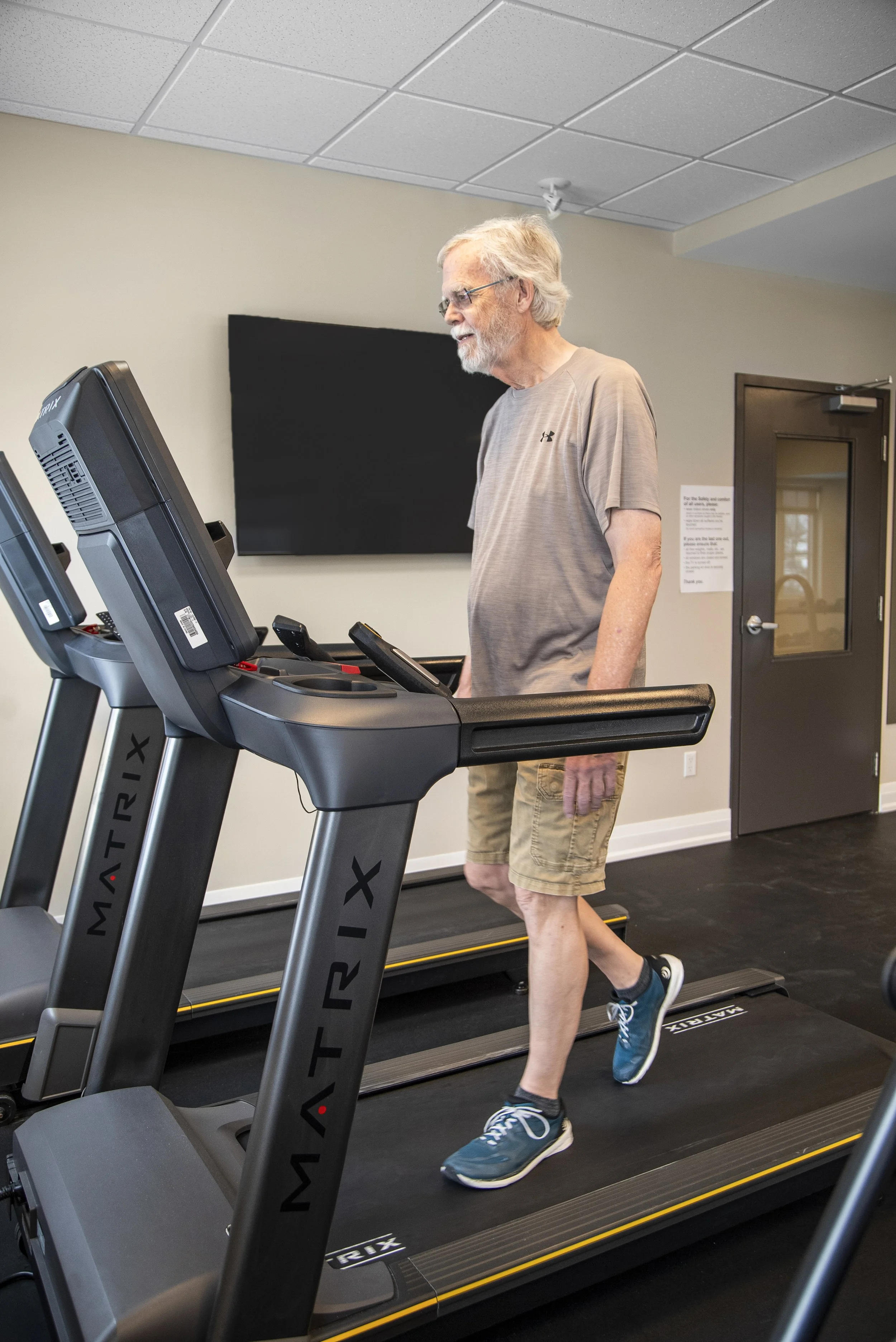 An elderly man with gray hair and a beard walking on a treadmill in a gym.