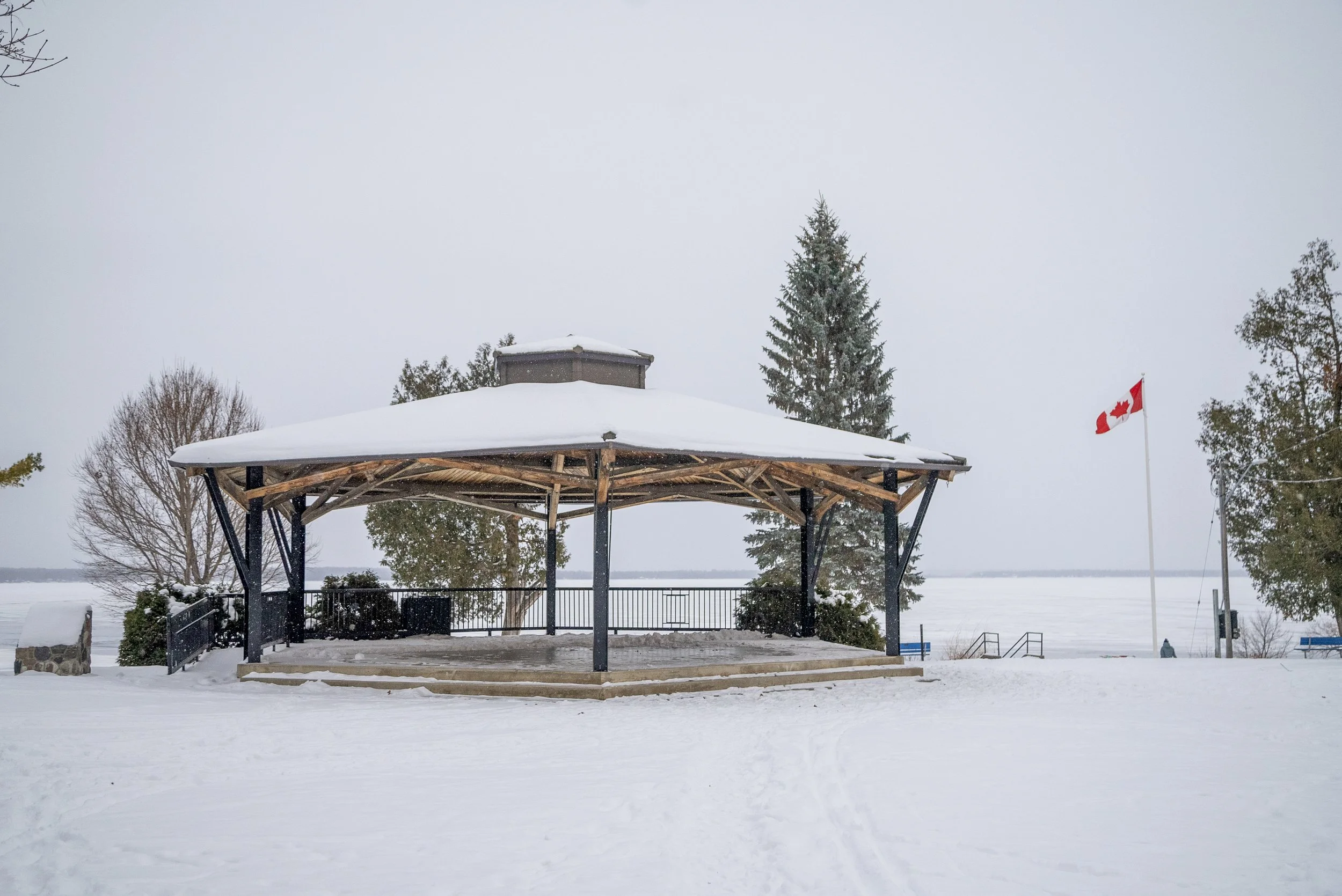 A snow-covered gazebo near a body of water with trees in the background and a Canadian flag on a flagpole.