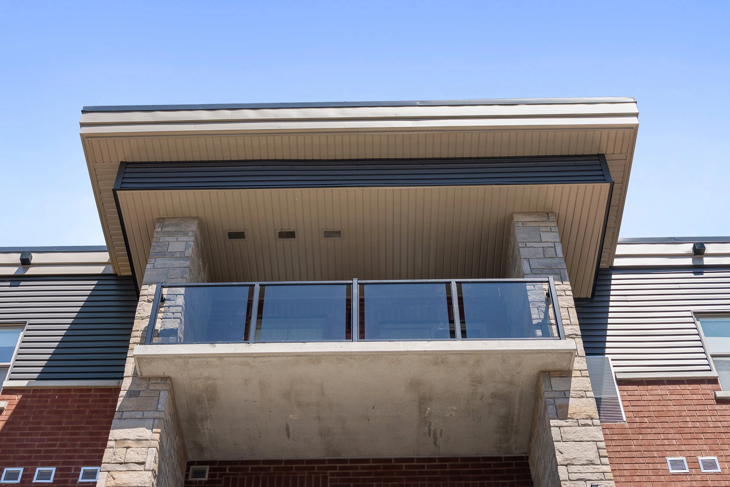 Exterior view of Sawmill Flats building featuring a covered balcony with stone pillars, dark siding, glass railing, and brick accents.