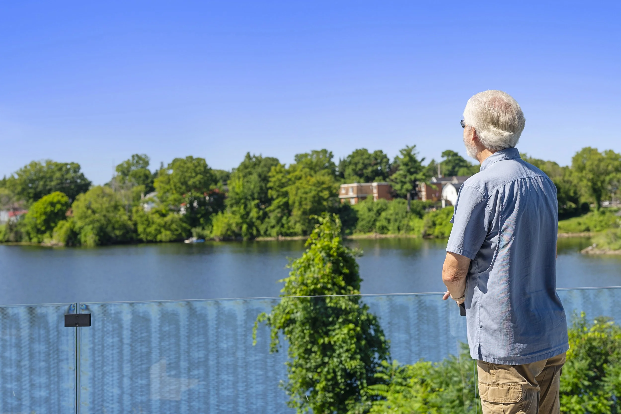 A man with gray hair standing out on a patio with glass railing, looking out over a wide river surrounded by trees.