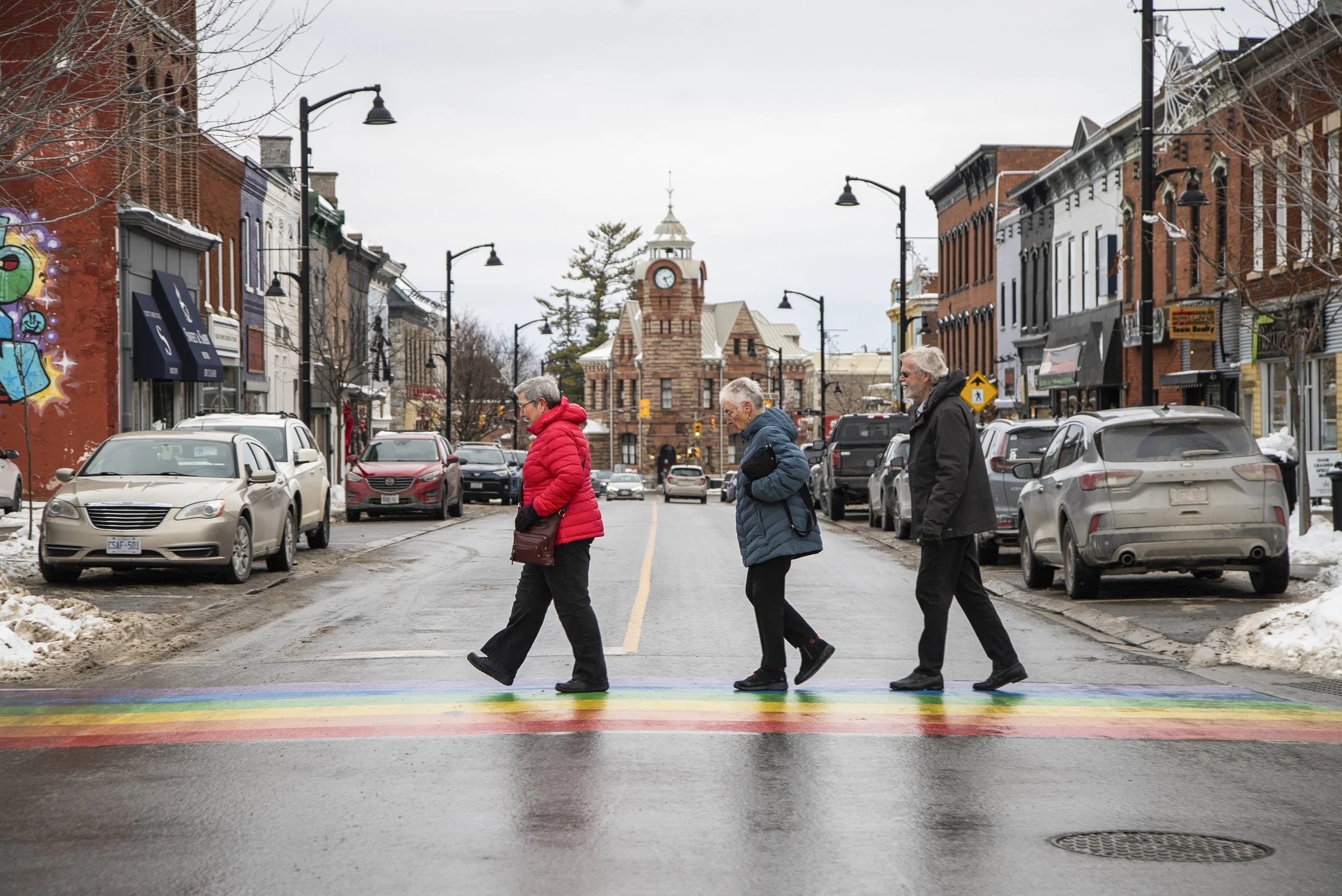 People crossing a rainbow-colored crosswalk on a city street with parked cars and historic-style buildings in the background.
