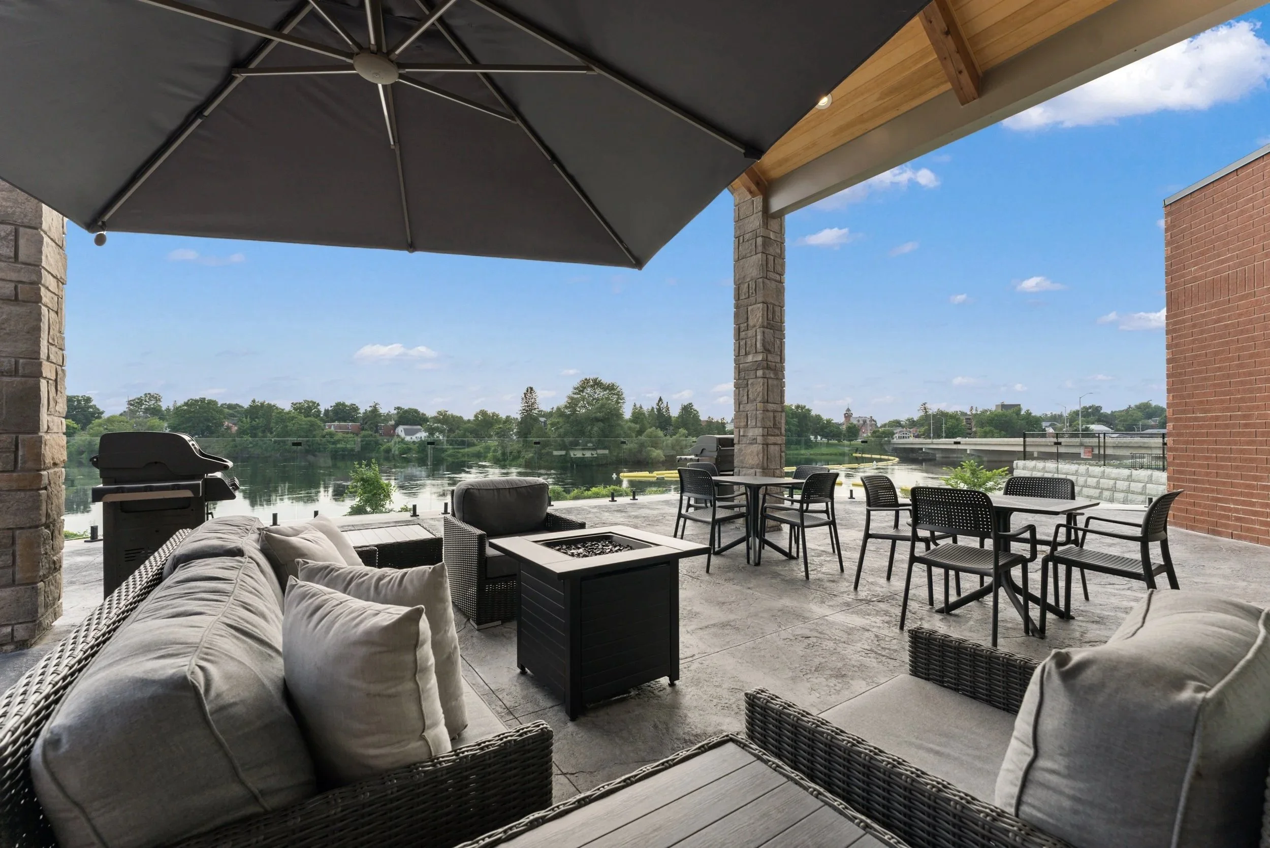 Outdoor patio overlooking a river, with cushioned seating, chairs, tables, a grill, and a large umbrella for shade.