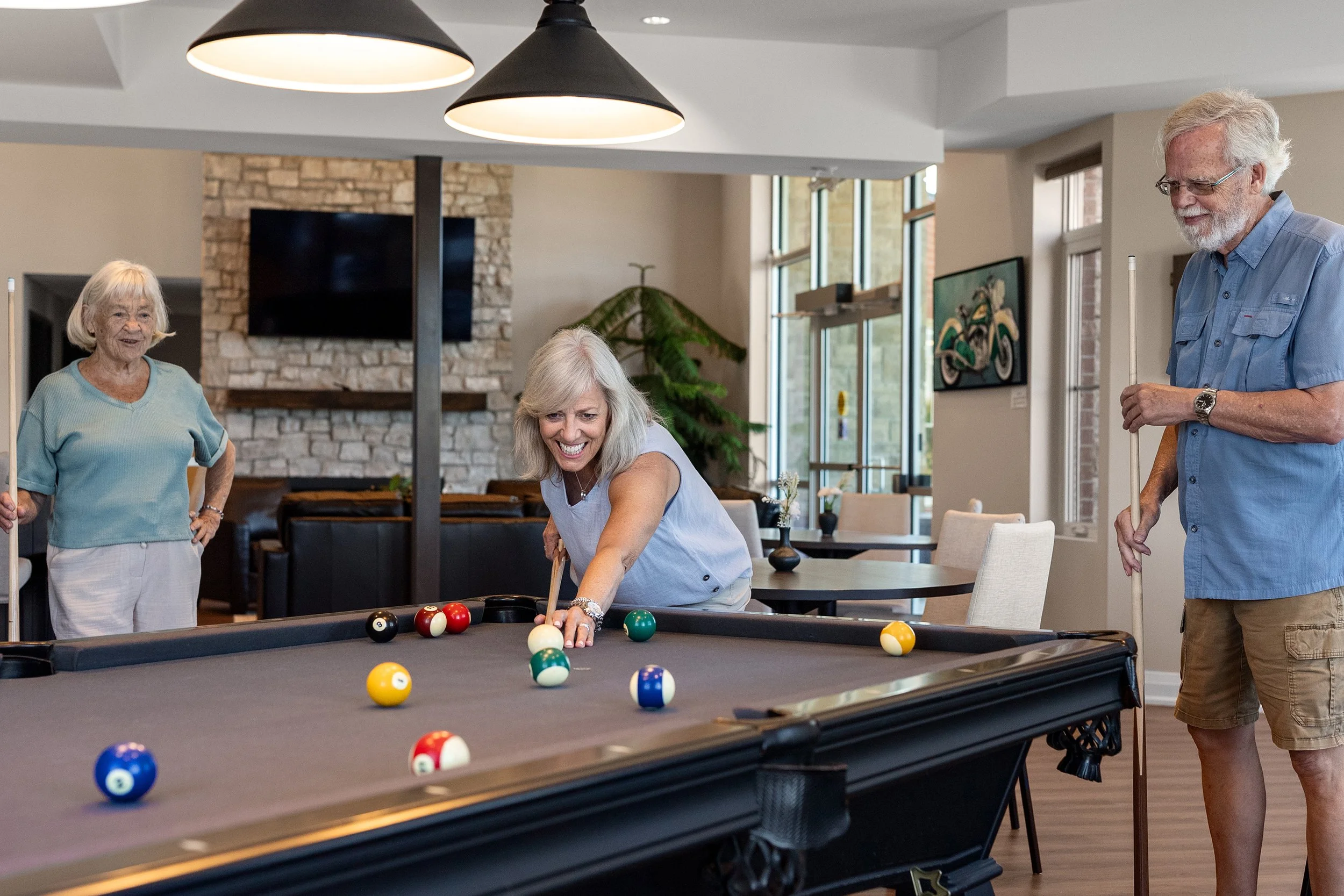 A woman leaning over to line up a pool shot while two friends watch and smile around a billiards table