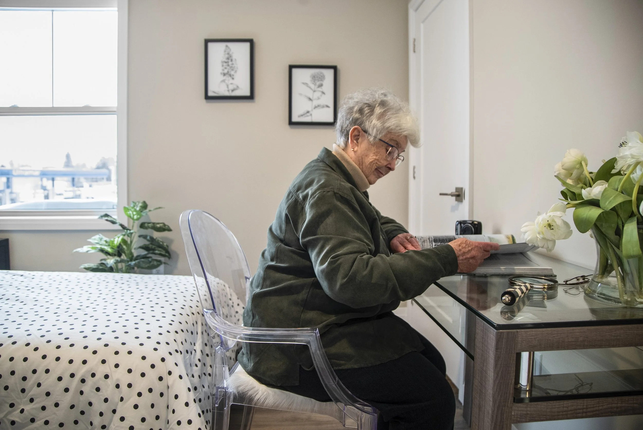 An elderly woman with gray hair and glasses sitting at a glass table, reading a newspaper, with a vase of white flowers, on a clear chair, in a bright room with a large window and framed black-and-white botanical prints.