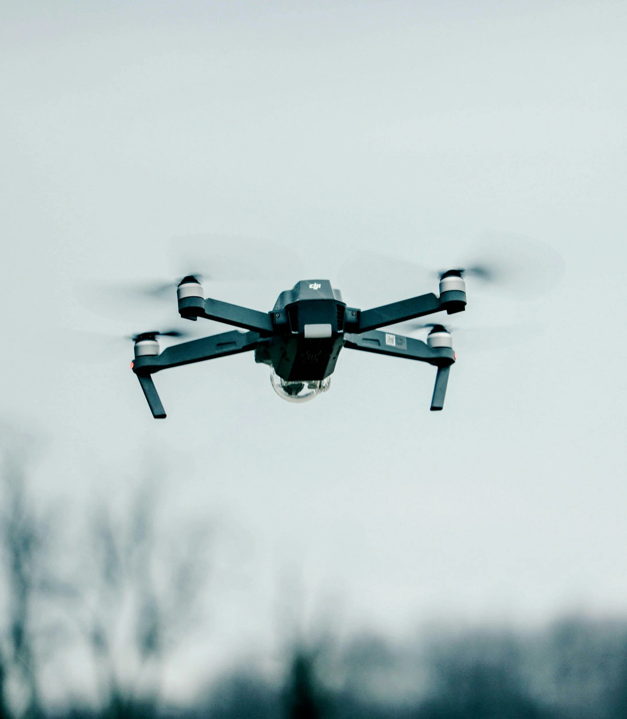 A drone flying against a cloudy sky with blurred foliage in the foreground.