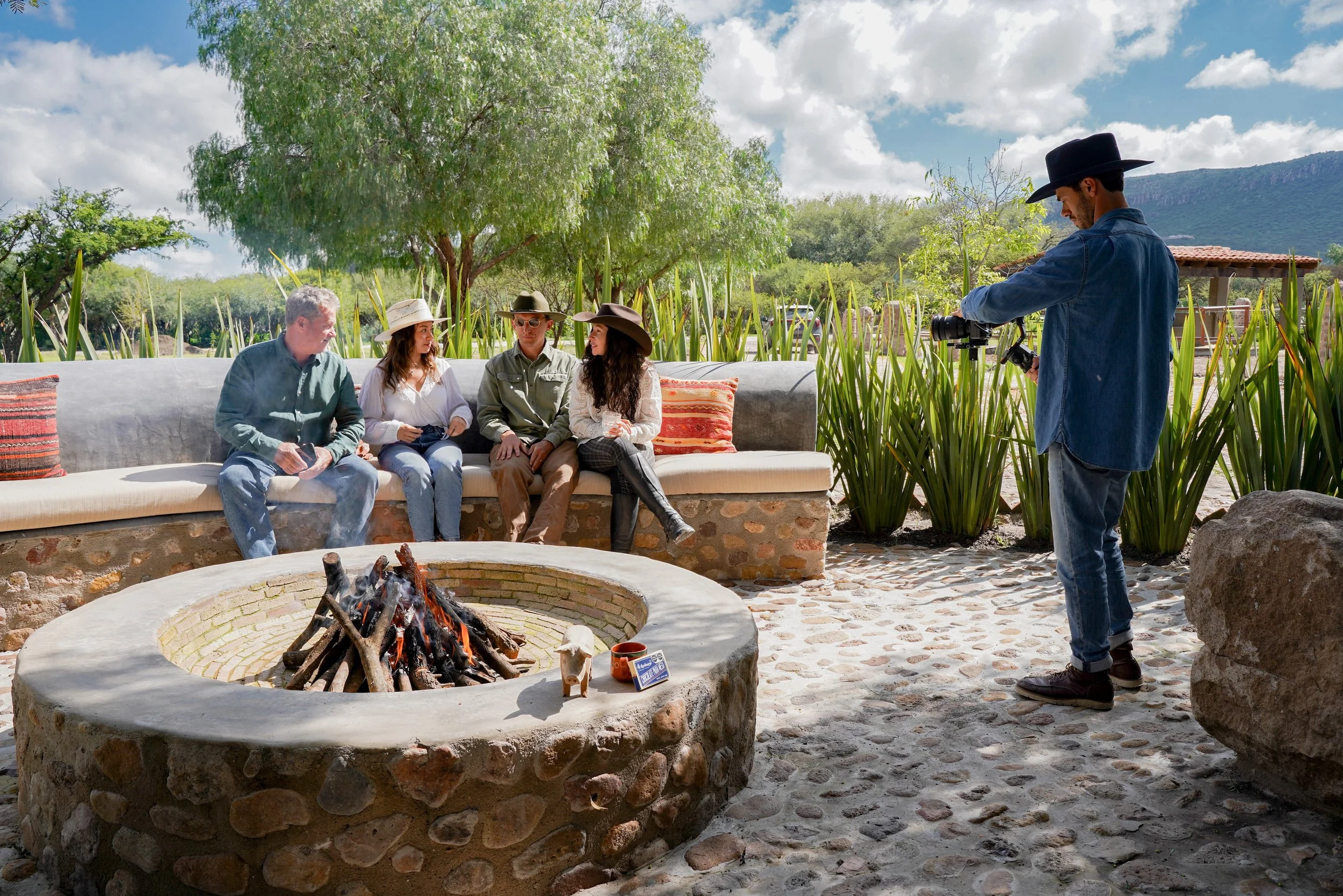 A group of four people sitting on a stone and concrete bench around a fire pit, being photographed by a man in cowboy attire with a camera. The setting is outdoors with lush green plants, trees, mountains in the background, and a partly cloudy sky.