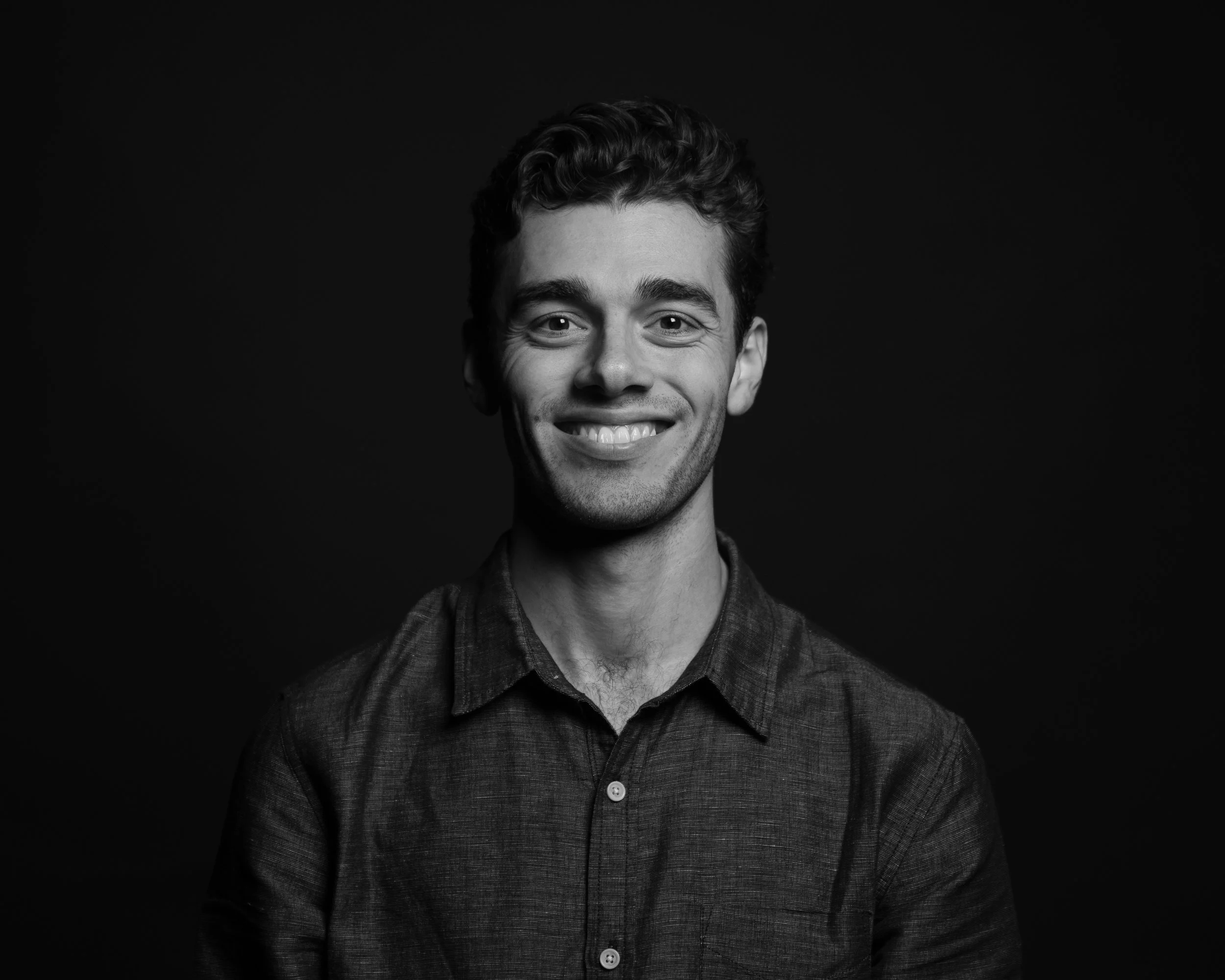 Black and white portrait of a smiling young man with wavy hair, wearing a button-up shirt, against a dark background.