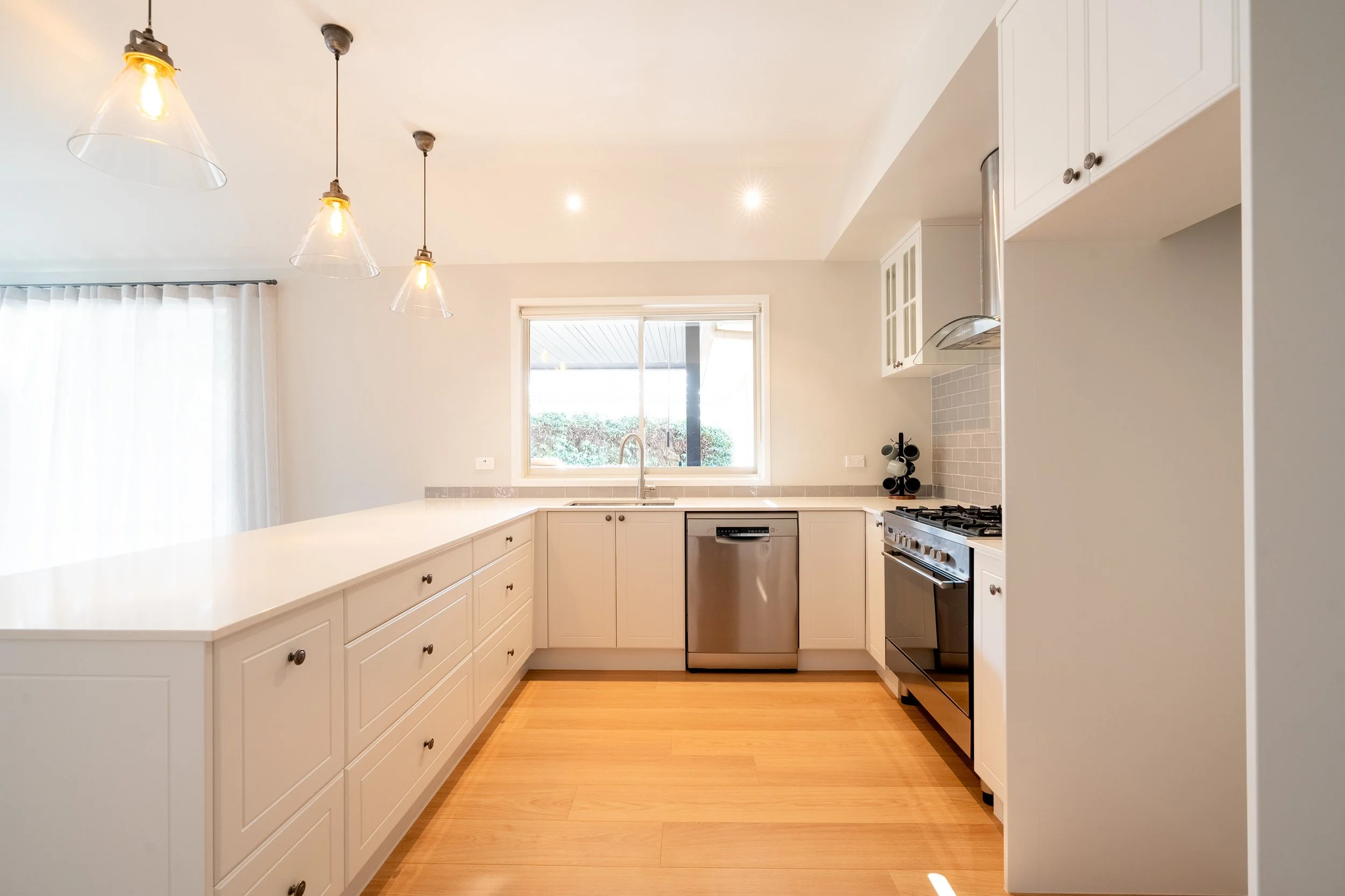 Modern kitchen with white cabinets, light wood flooring, stainless steel dishwasher, and stove. Kitchen window above the sink, pendant lights hanging from the ceiling, and a door with white curtains on the left.