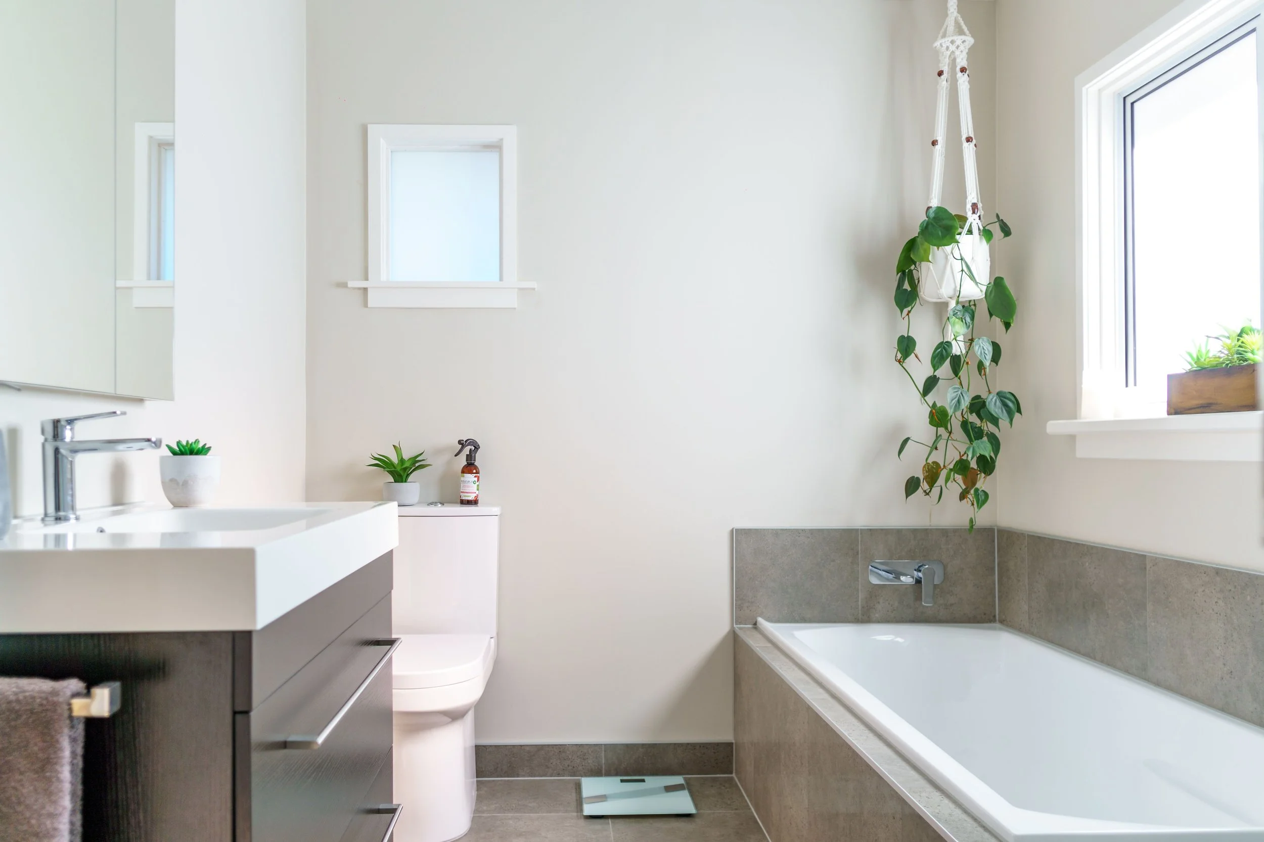 Modern bathroom with white walls, a bathtub next to a window with plants, a small mirror on the wall, a dark cabinet with a towel, a white sink with a soap dispenser, two small potted plants, a scale, and a hanging plant in a white macrame planter.