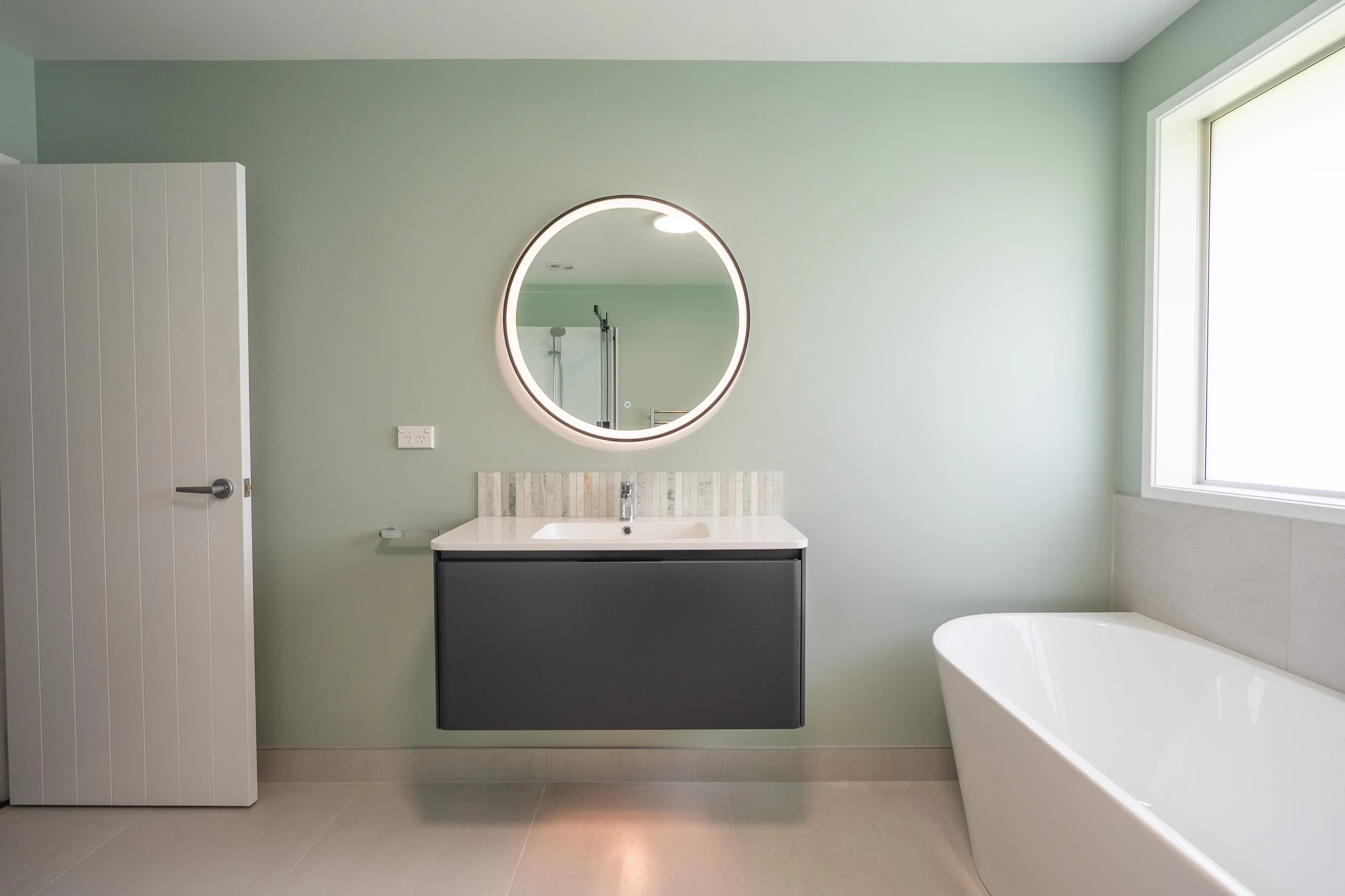 Modern bathroom with a green wall, a floating vanity with a white countertop and mirror, a bathtub, and a window providing natural light.