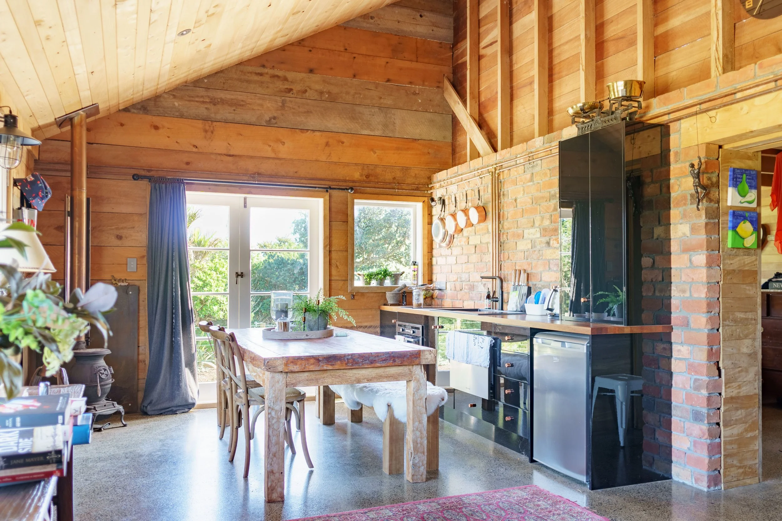 Open-concept kitchen and dining area with wood-paneled ceiling, brick and wooden wall, dining table, chairs, and large windows with a view of greenery.