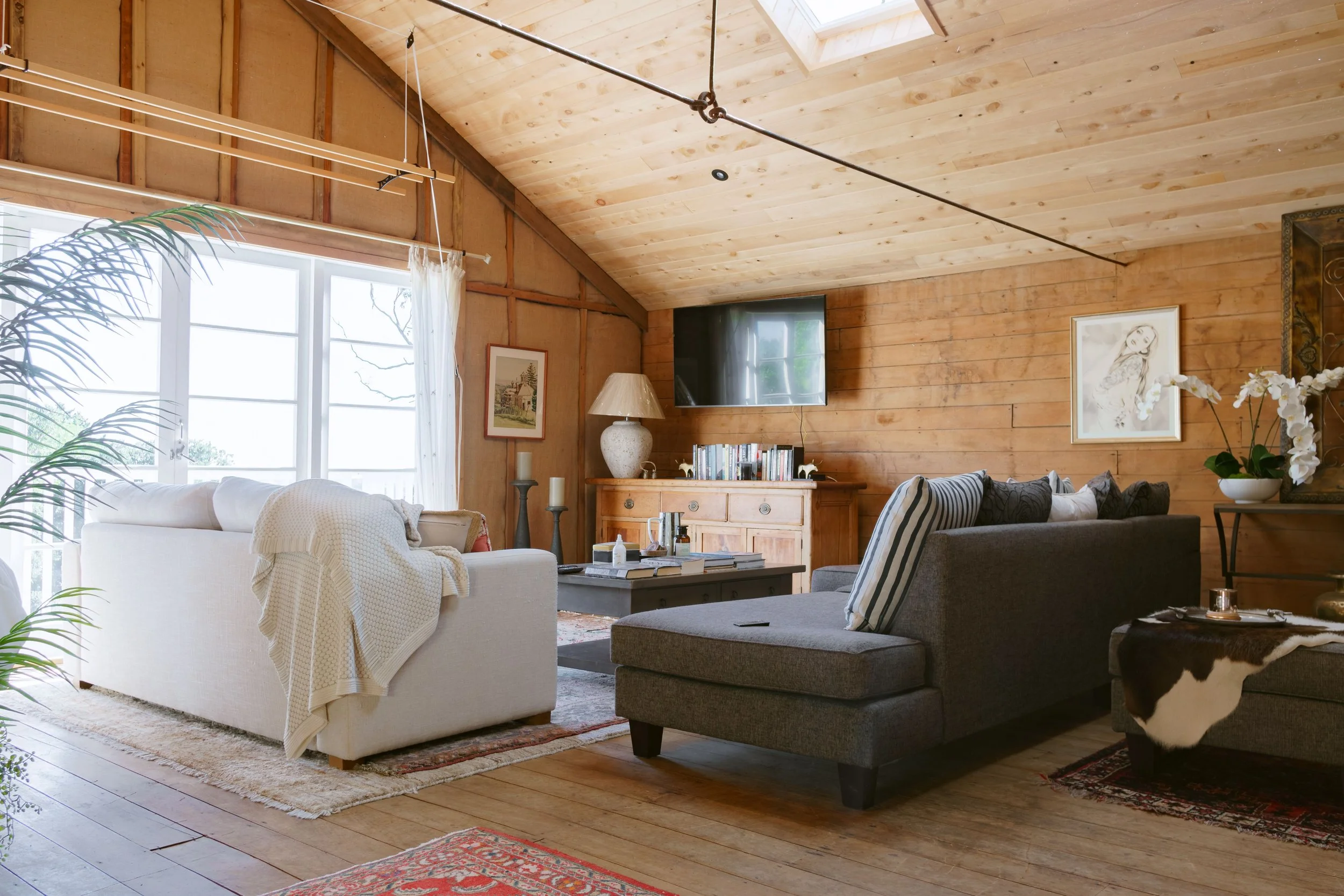 Living room with wooden walls and ceiling, large window with curtains, two sofas (one white and one gray), coffee table, TV mounted on wall, bookshelf, decorative items, and artwork.