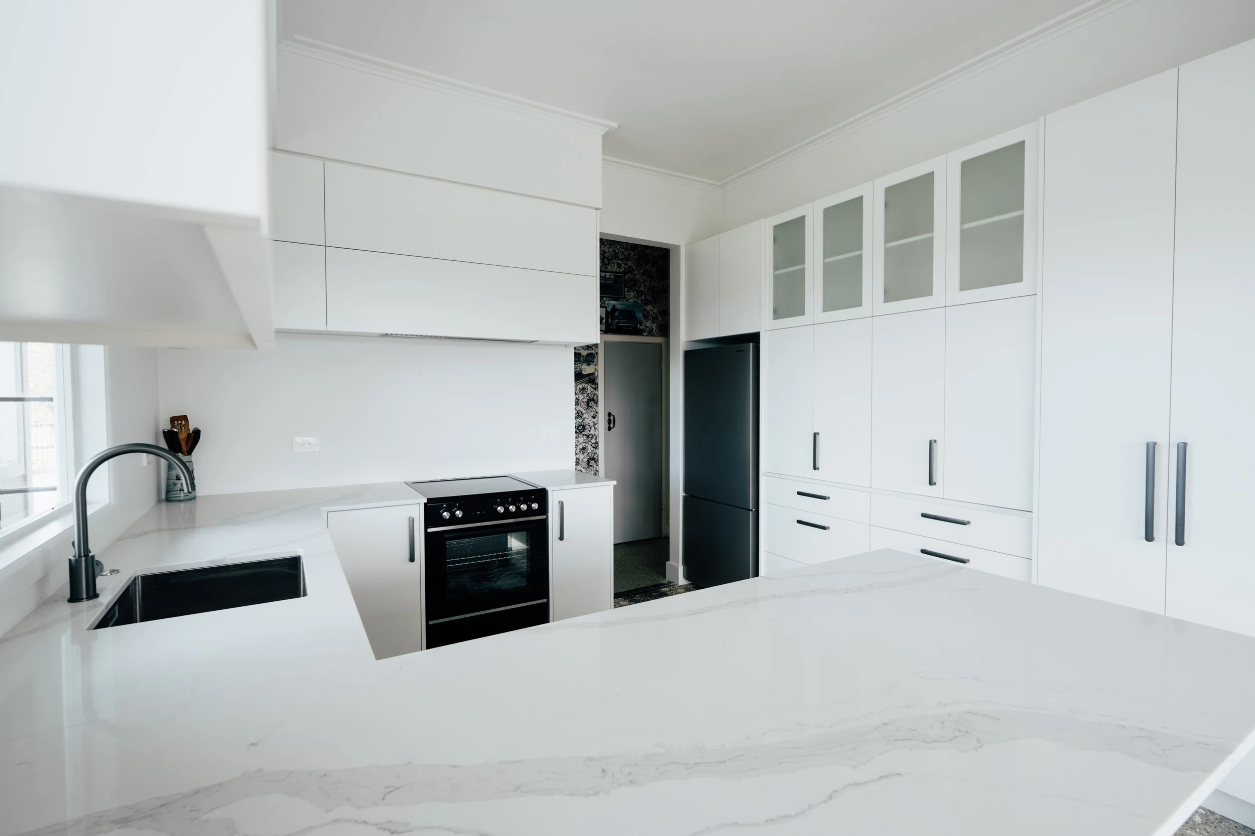 Modern white kitchen with black appliances, including a refrigerator and oven, with marble countertops and glass-front cabinets.