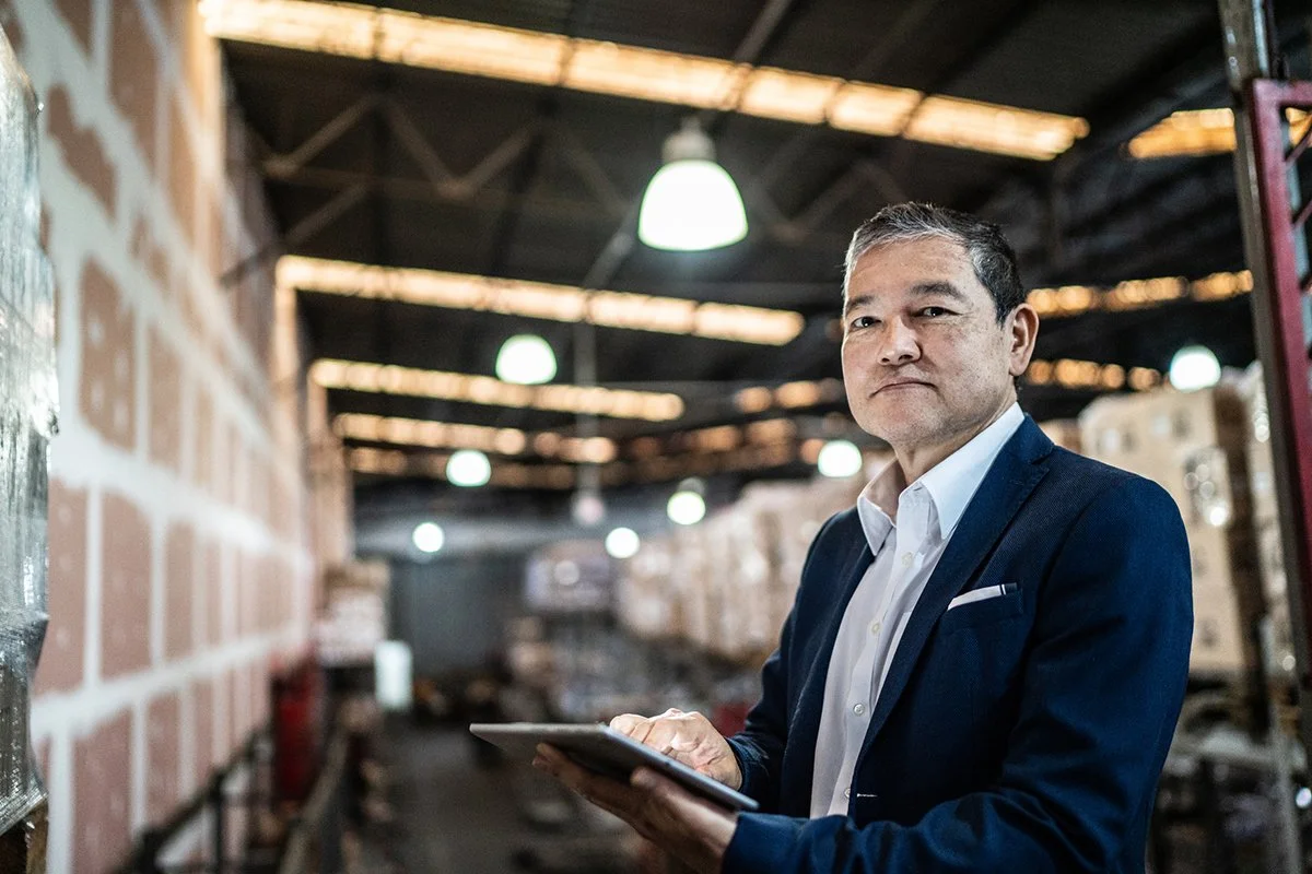 A mature male business owner in a blue blazer and white shirt looking at an ipad in his warehouse