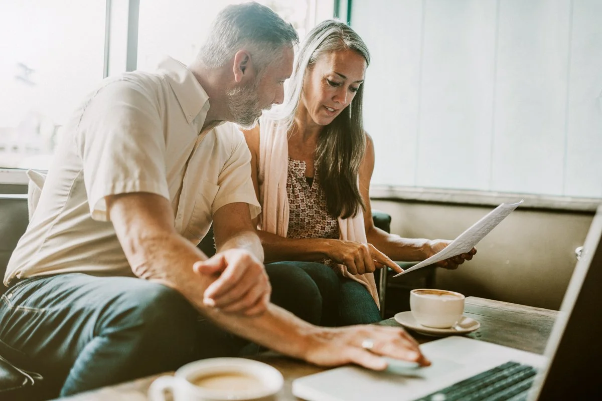 A middle-aged man and woman sitting together at a table in a cafe, looking at a laptop and a document, with coffee cups in front of them.