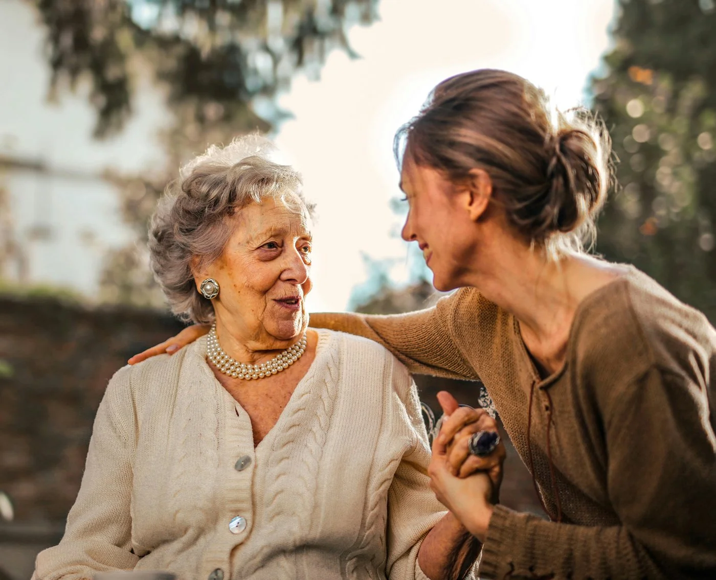 A younger blonde woman holding the hand of a seated, much older woman, wearing nice clothes and pearls