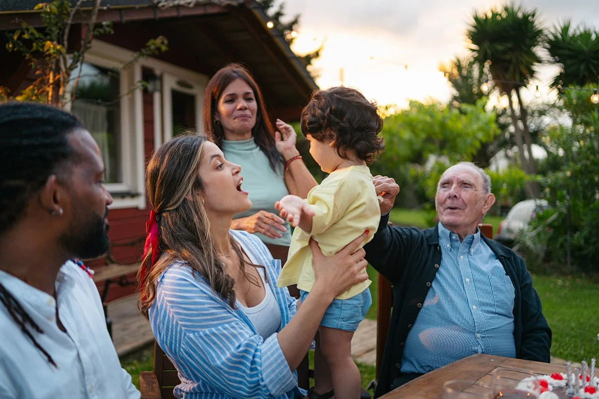 A mixed-race family seated at an oudoor table, including a husband and wife, older sister, toddler boy, and elderly grandfather. 