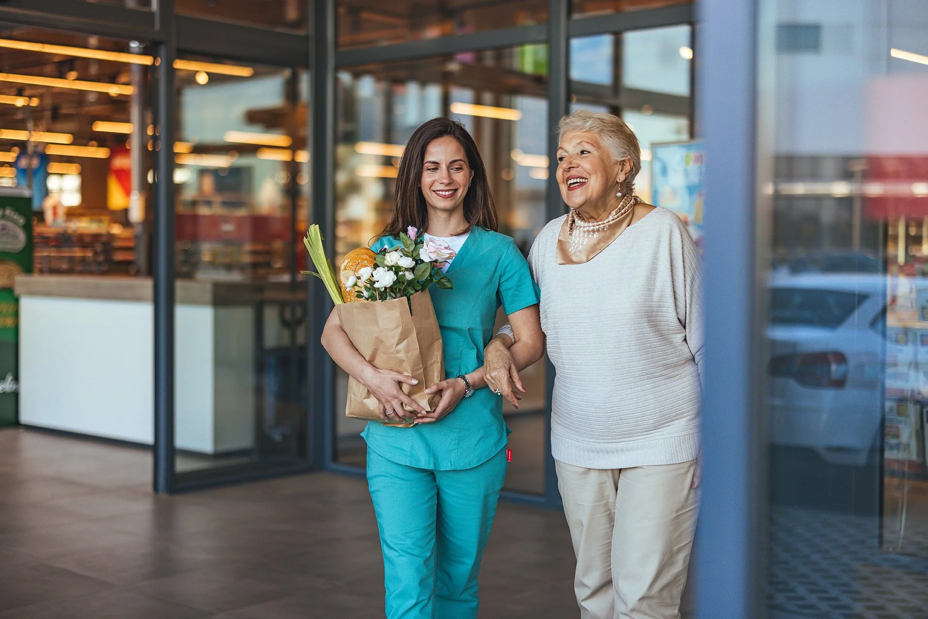A young woman in blue scrubs holding a paper bag with flowers and groceries, walking with an elderly woman outside a store, both smiling and engaging in conversation.