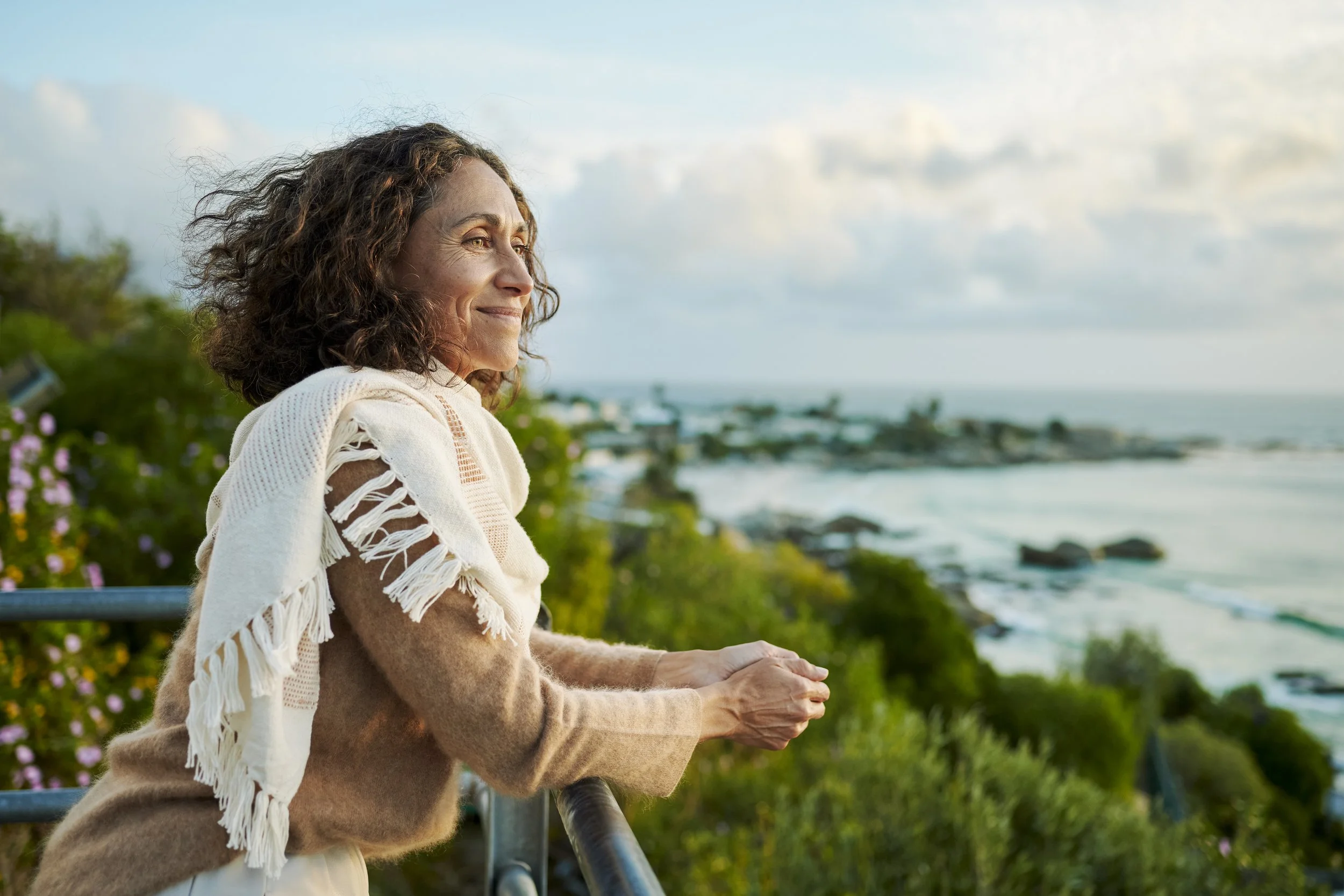 A middle-aged woman with brown curly hair, wearing a light sweater and scarf, standing at a railing looking out over the beach