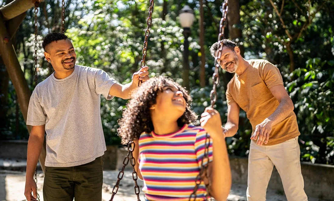 A mature, mixed-race gay couple pushing their tween daugher on a swing at the park