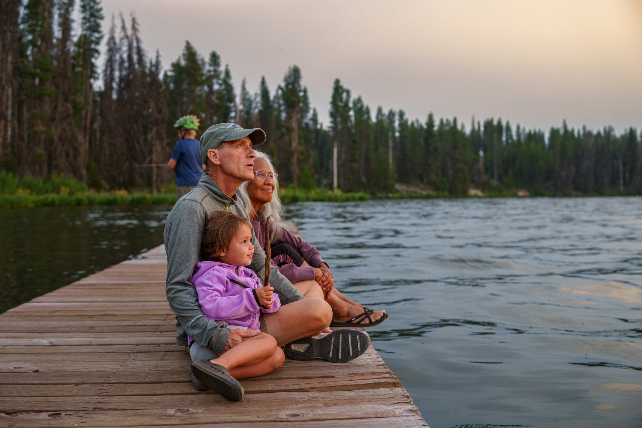 Mixed-race grandparents sitting on the dock of a lake with their two grandchildren, with trees in the background, looking off into the sky